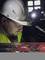 A construction worker wearing a hard hat and headlamp, looking down at his hands holding a tool. Background dark, focus on the work.