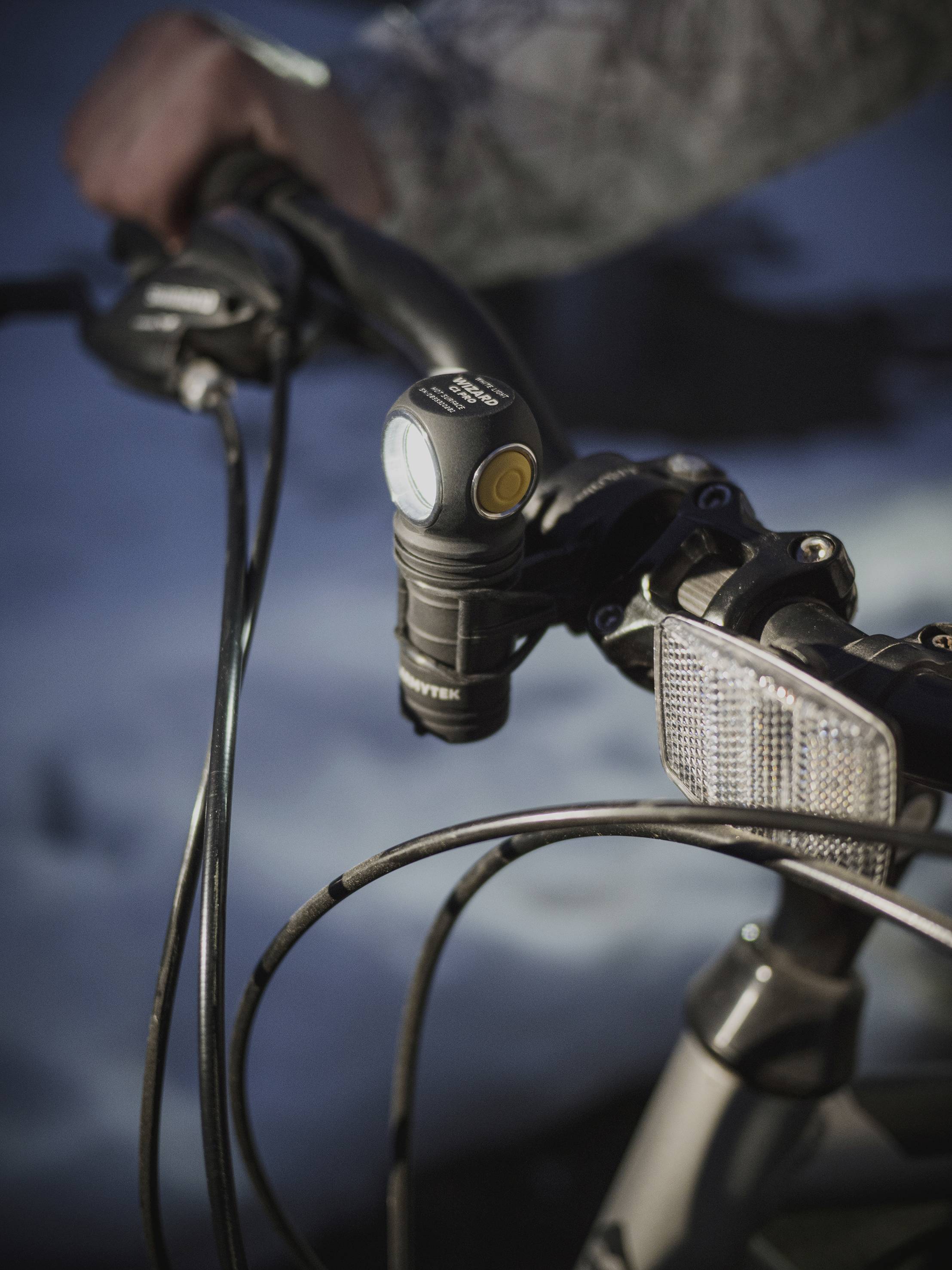 A hand grasps the handlebars of a bicycle, equipped with a lamp and reflector. The background is slightly blurred and dark.