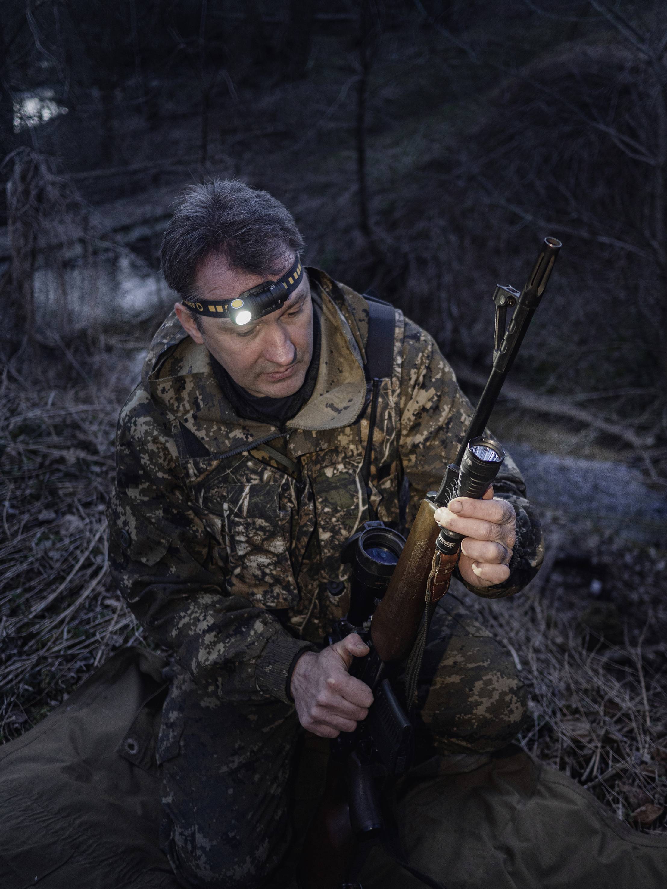 A hunter in camouflage clothing sits in the woods and inspects his rifle. It is dark, and he is wearing a head torch for light.