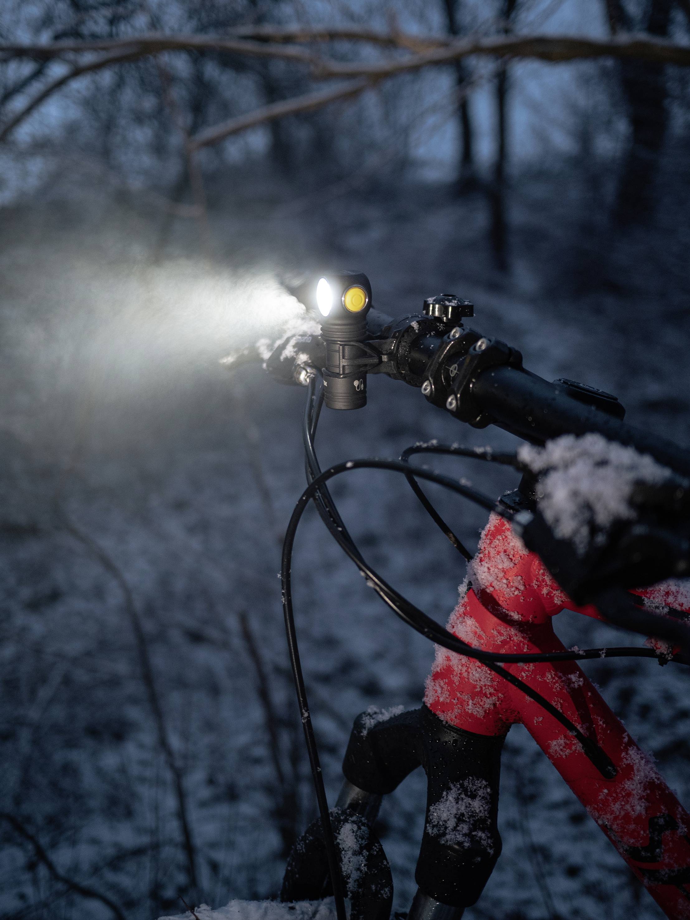 A bicycle in falling snow with its front light switched on. The background shows a blurred forest in the darkness.
