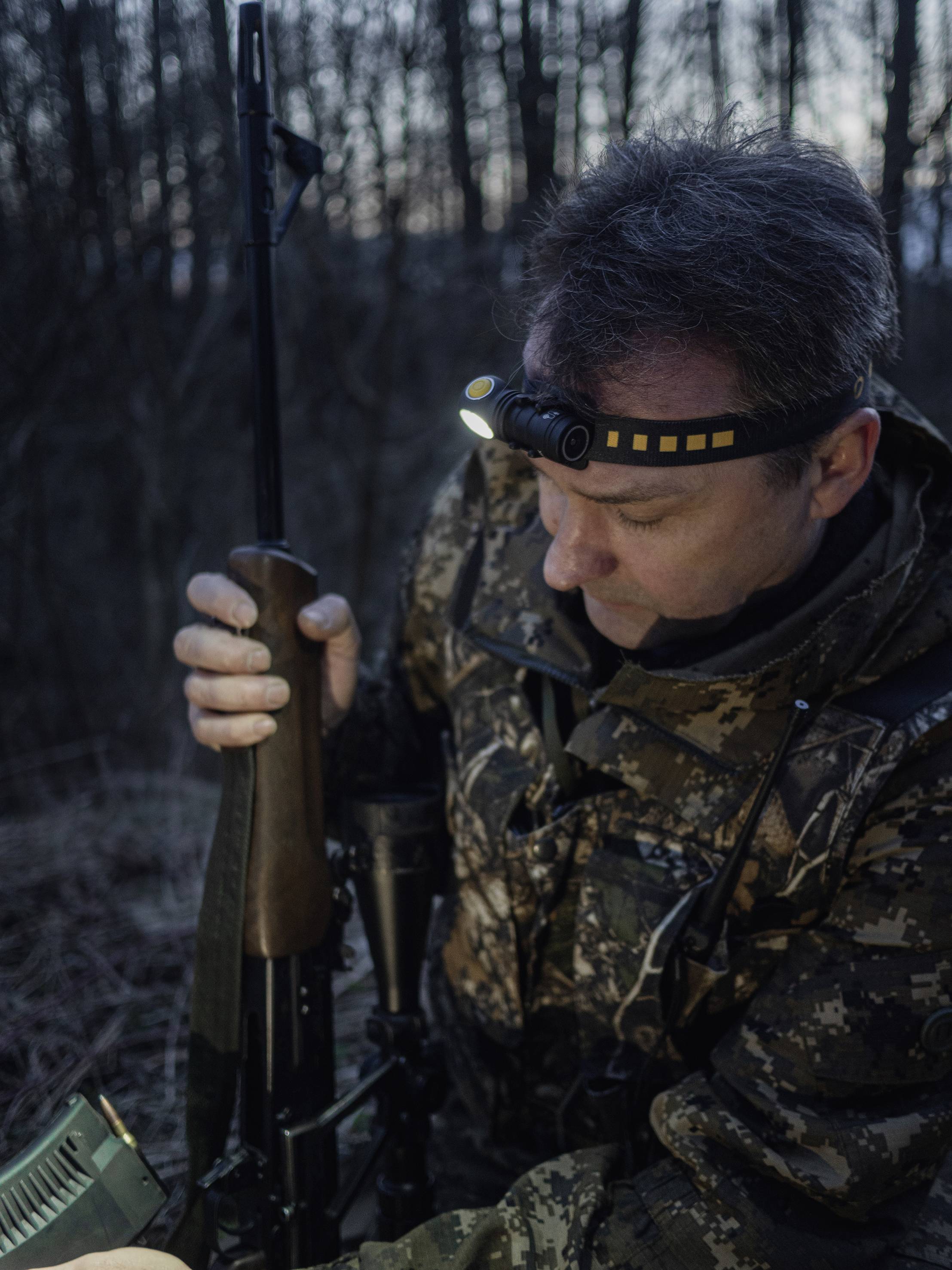 A hunter in camouflage stands in a forest at dusk. He is holding a rifle and wearing a head torch that illuminates his path.