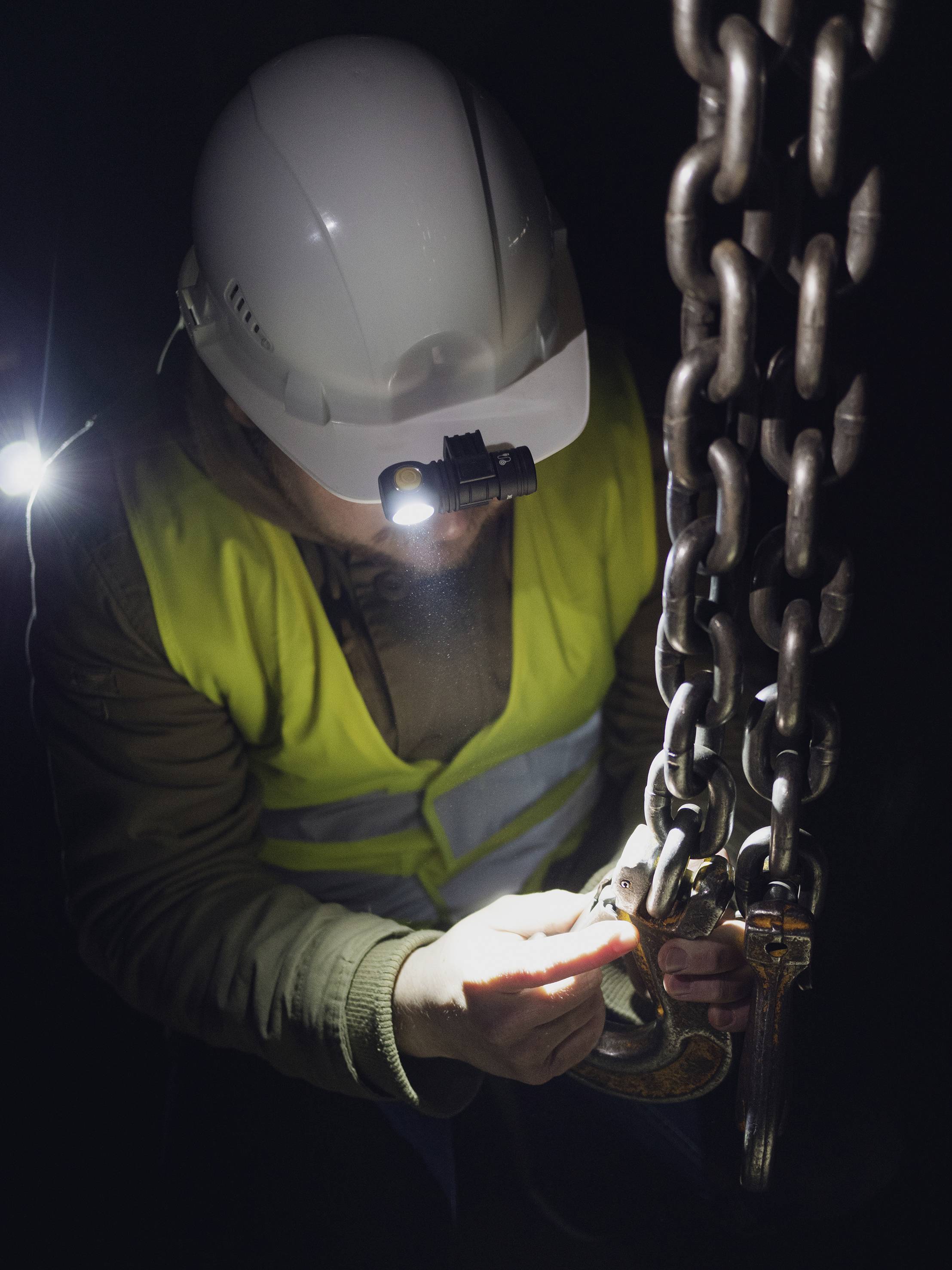 A person wearing a hard hat and high-visibility vest is inspecting a chain with a torch in a dimly lit work environment.