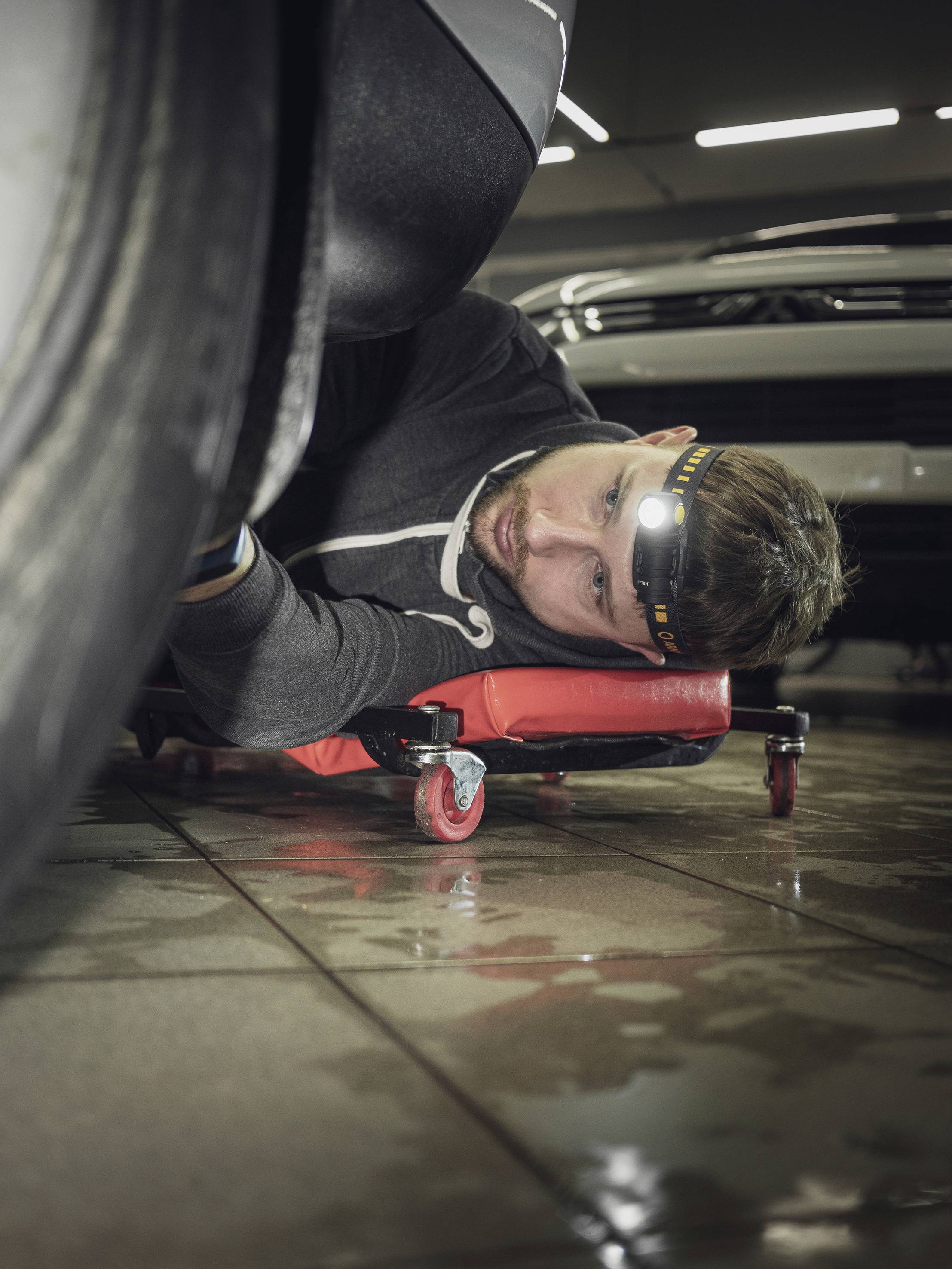 A man is lying on a skateboard underneath a car and working with a head torch in a workshop.