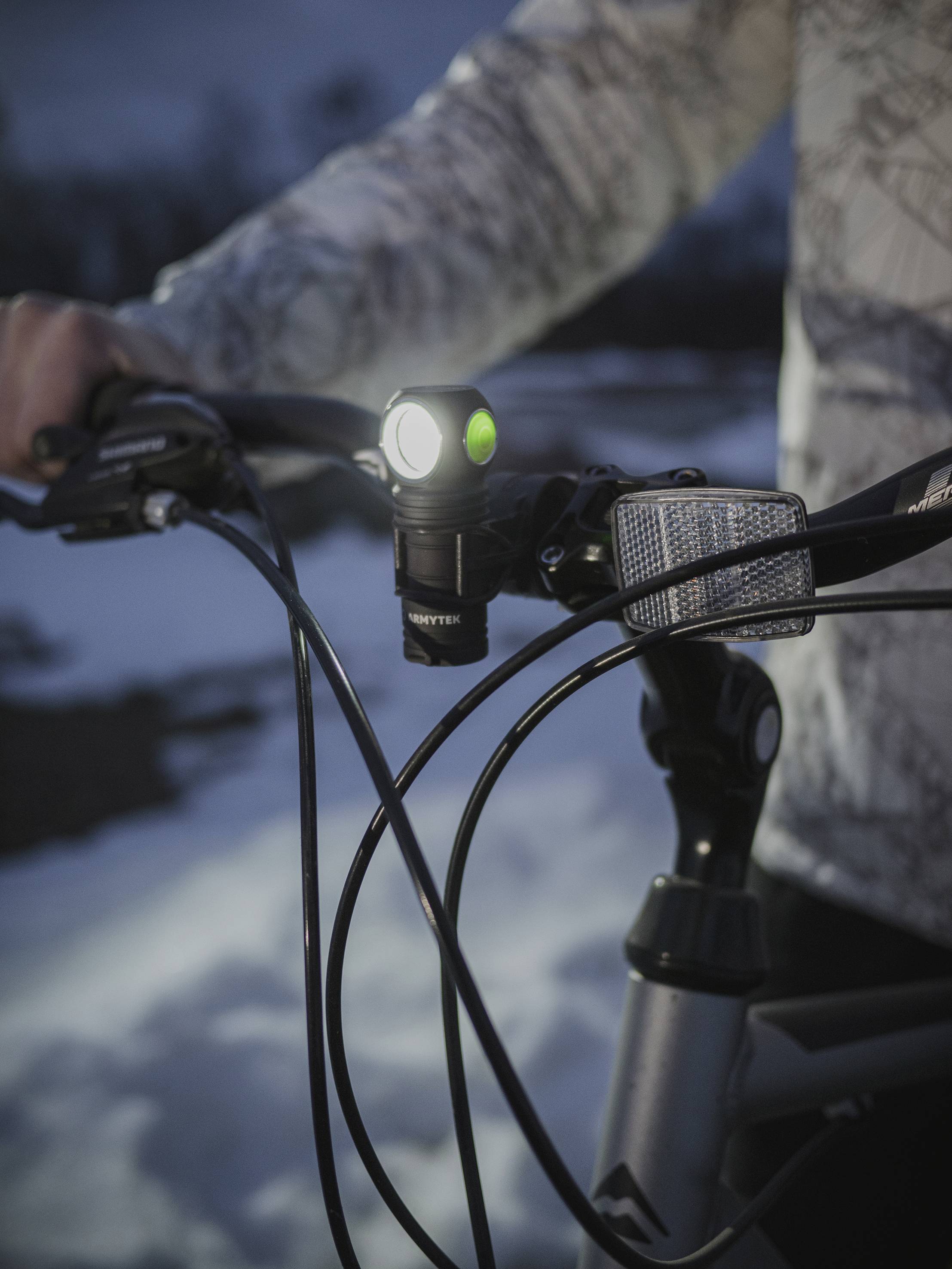 A bicycle handlebar in the snow is being held by a person. A bright front light is switched on, illuminating the path.