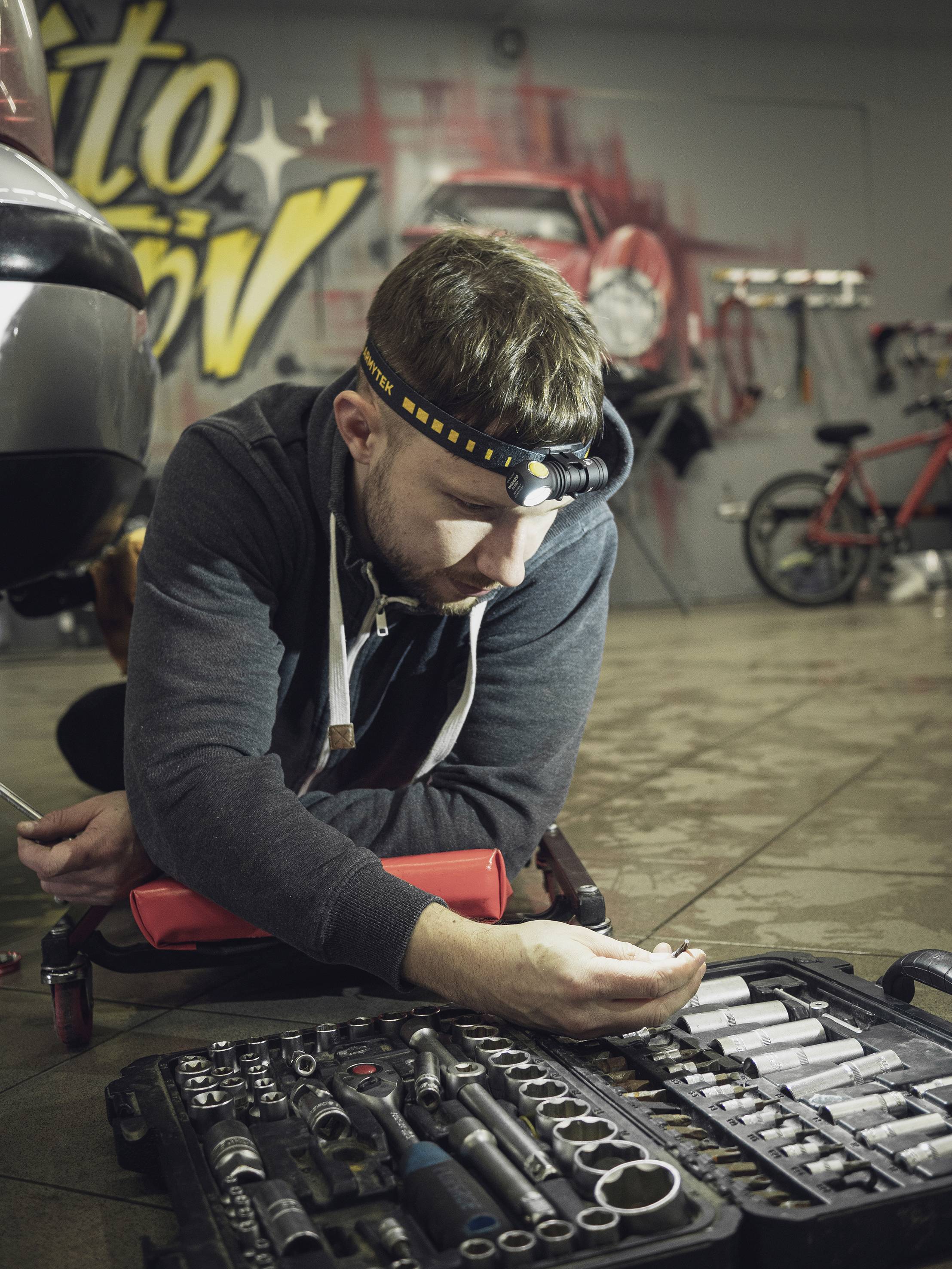 Man wearing a head torch repairs a car in a workshop; a toolbox lies in front of him. Graffiti and a red bicycle are in the background.