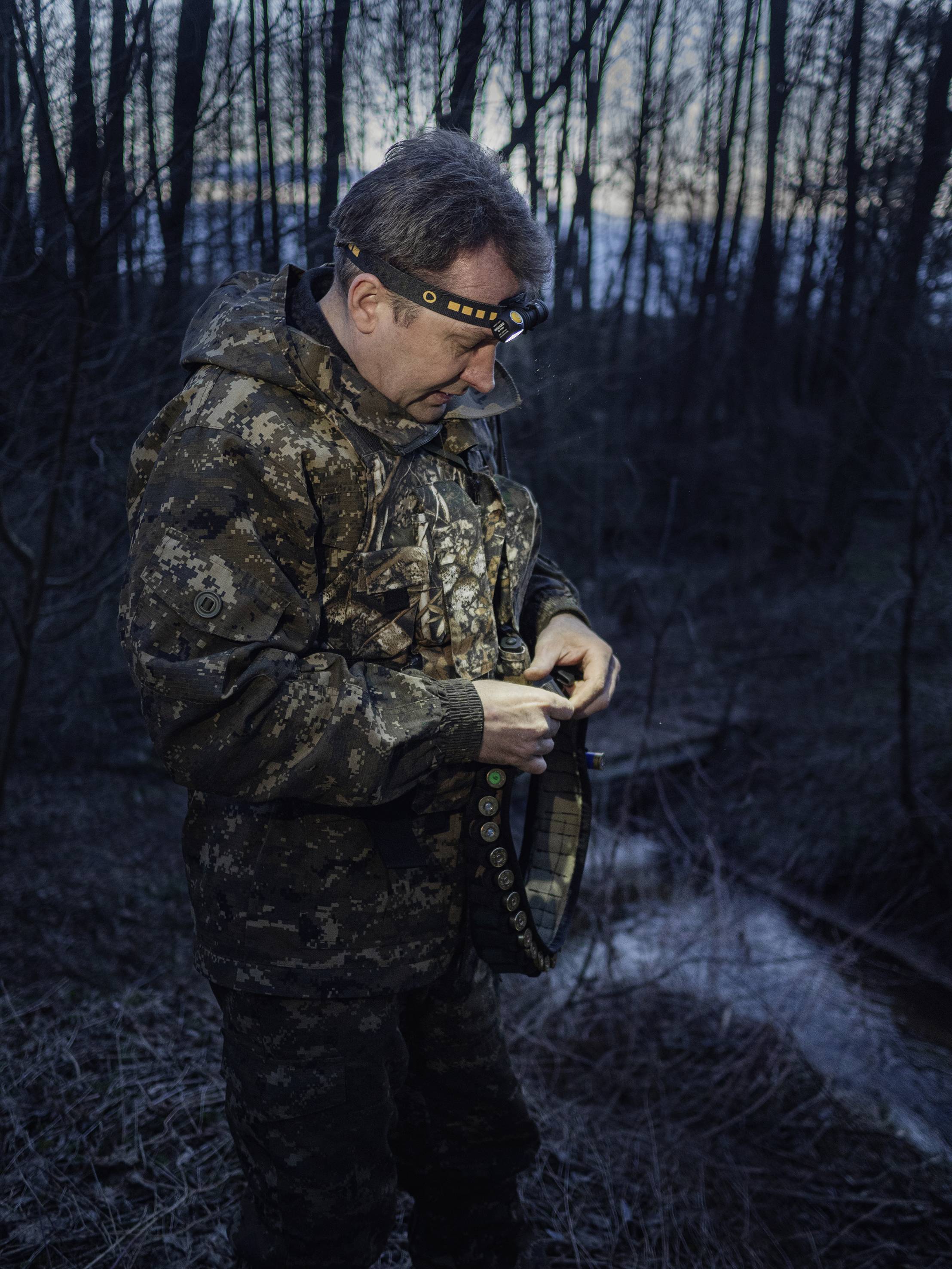 A man in camouflage clothing stands in the woods at dusk. He is wearing a head torch and holding a penknife. Trees form the background.