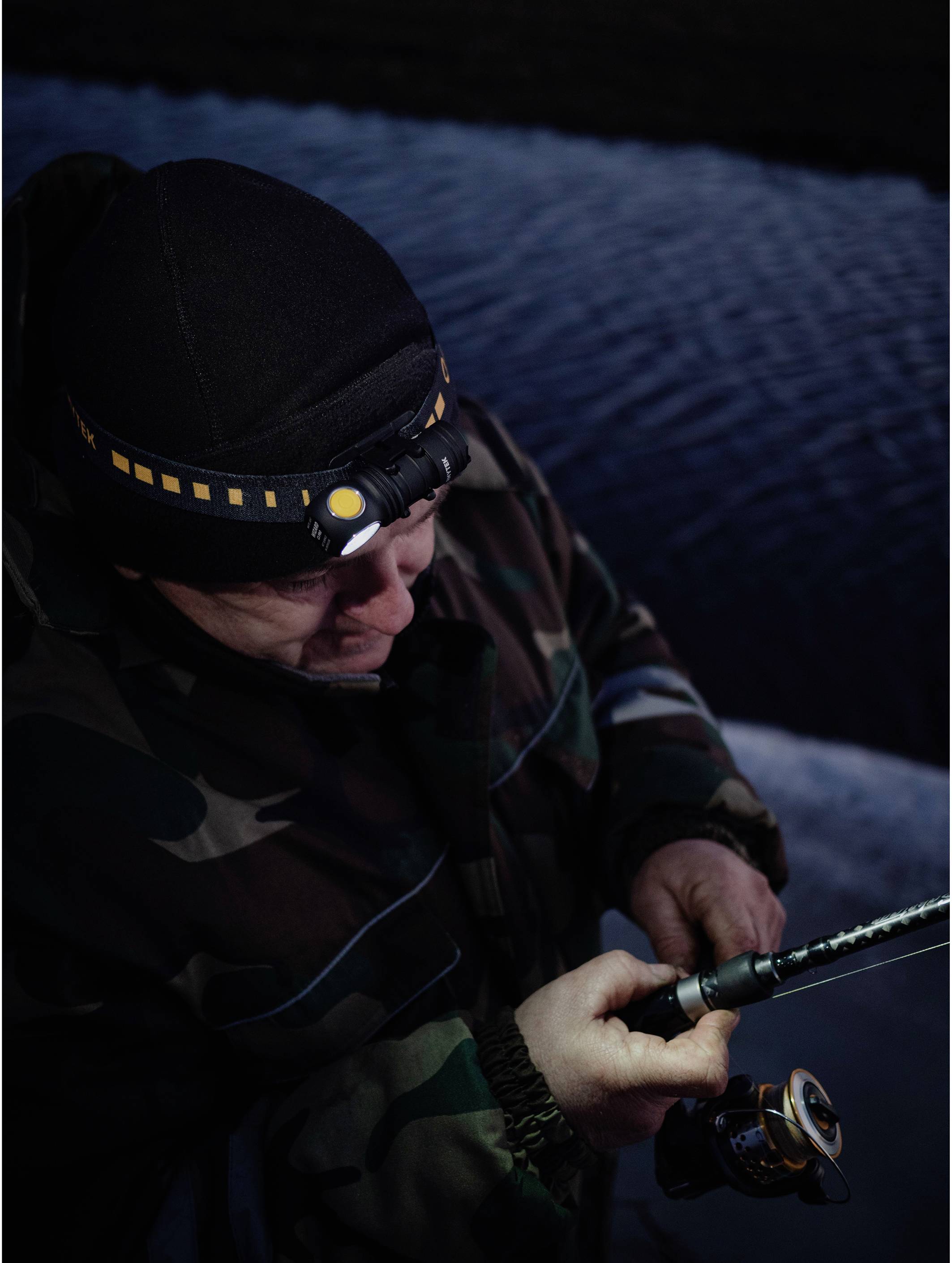 A person is fishing at night by the water, wearing camouflage clothing and a head torch.