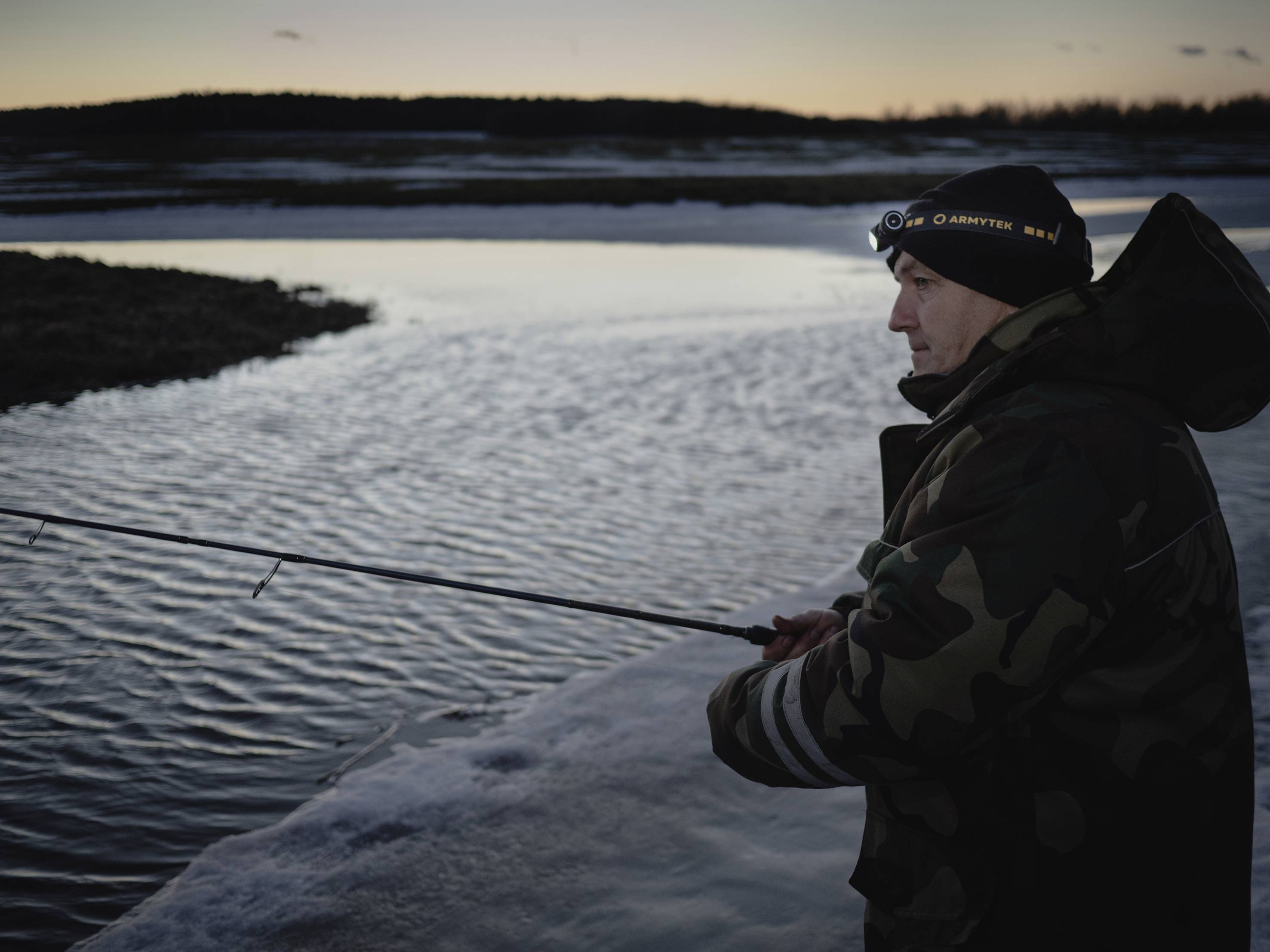 A person in winter clothing is fishing on the bank of a frozen river at dusk.