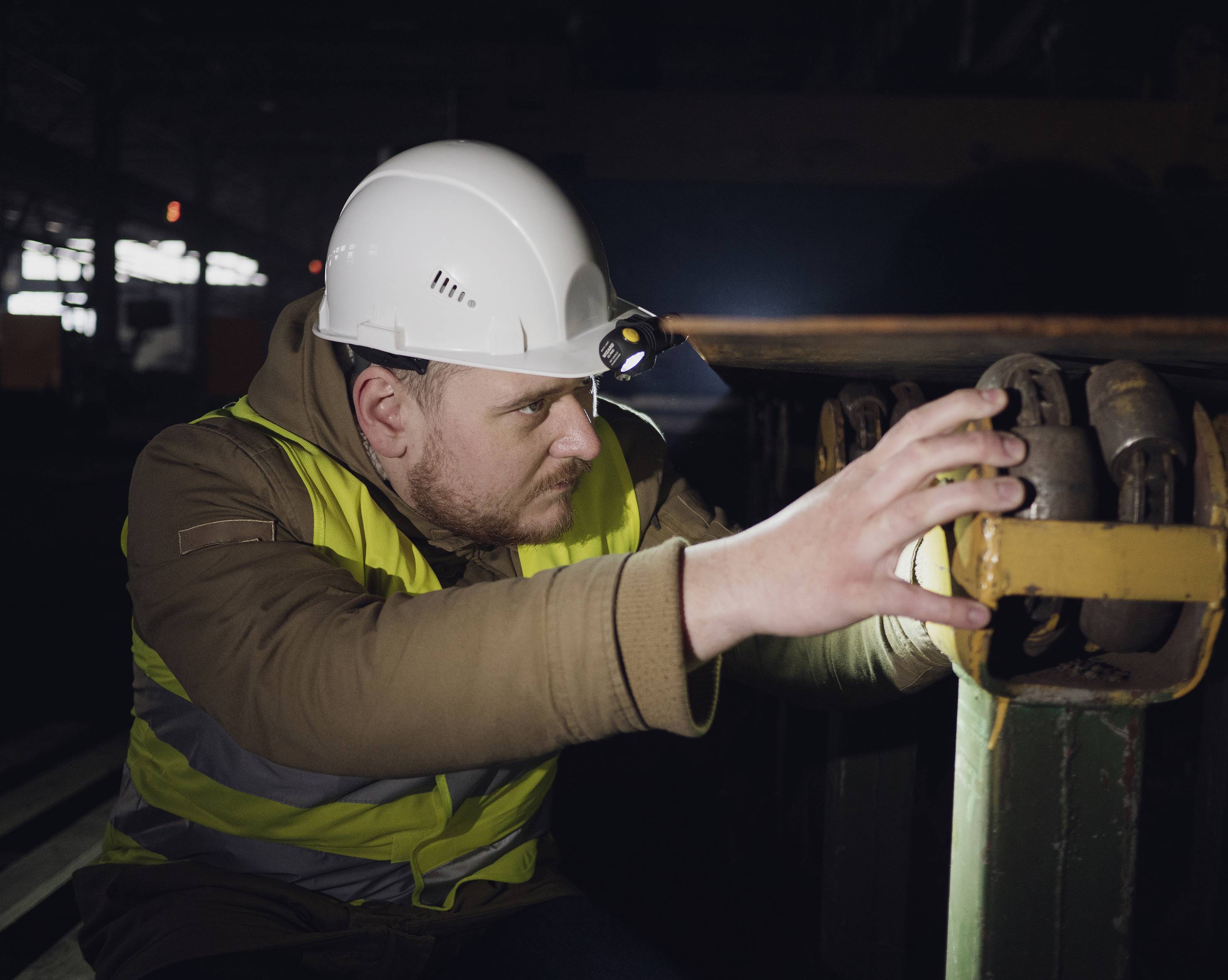 A man wearing a hard hat and high-visibility vest carefully inspects industrial rolls or wheels in a factory environment.