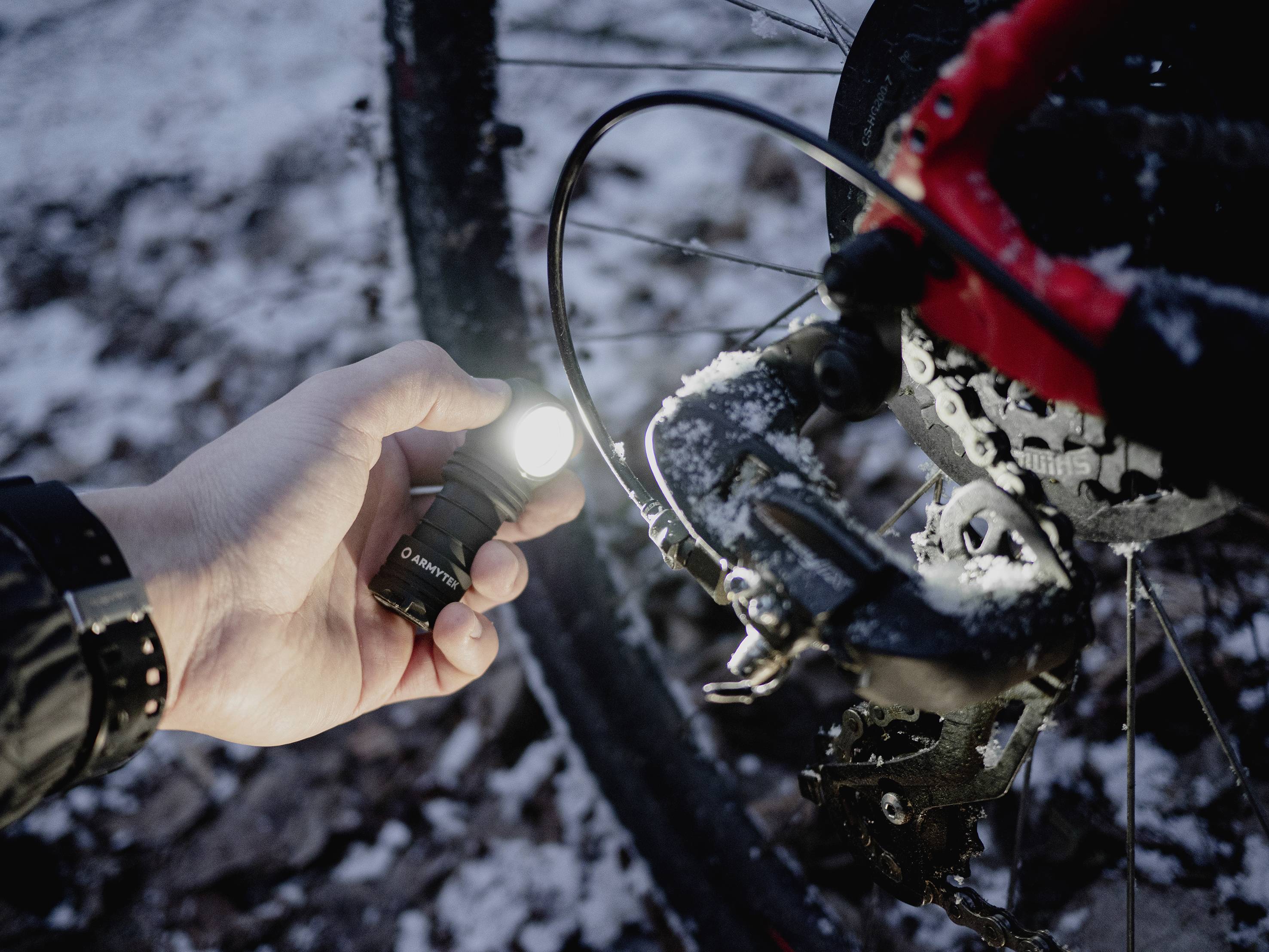 A hand holds a glowing torch and illuminates a bicycle chain in the snow. The scene depicts maintenance under wintry conditions.