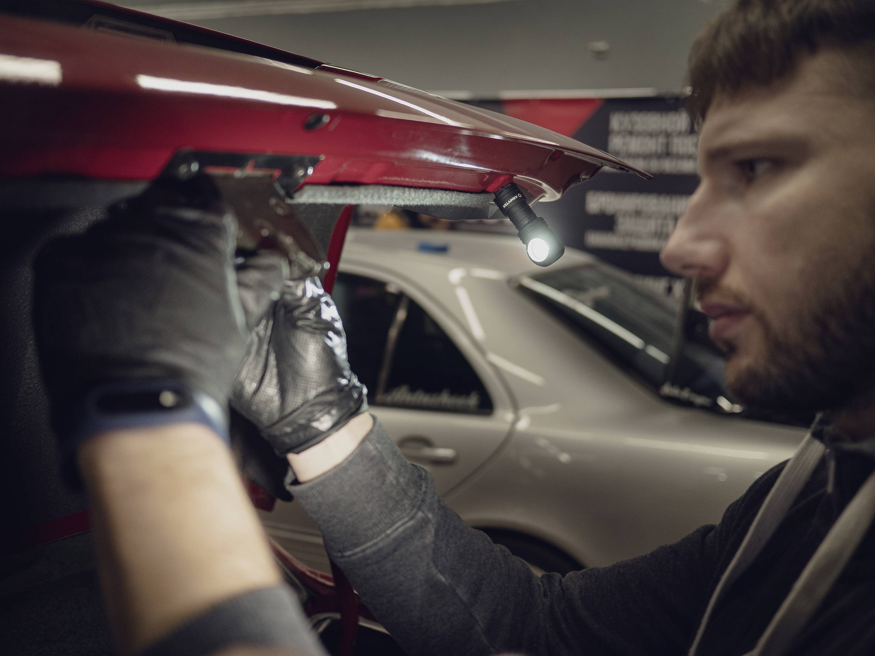 A man is repairing or inspecting the underside of a red car roof in a workshop. A silver car is standing in the background.