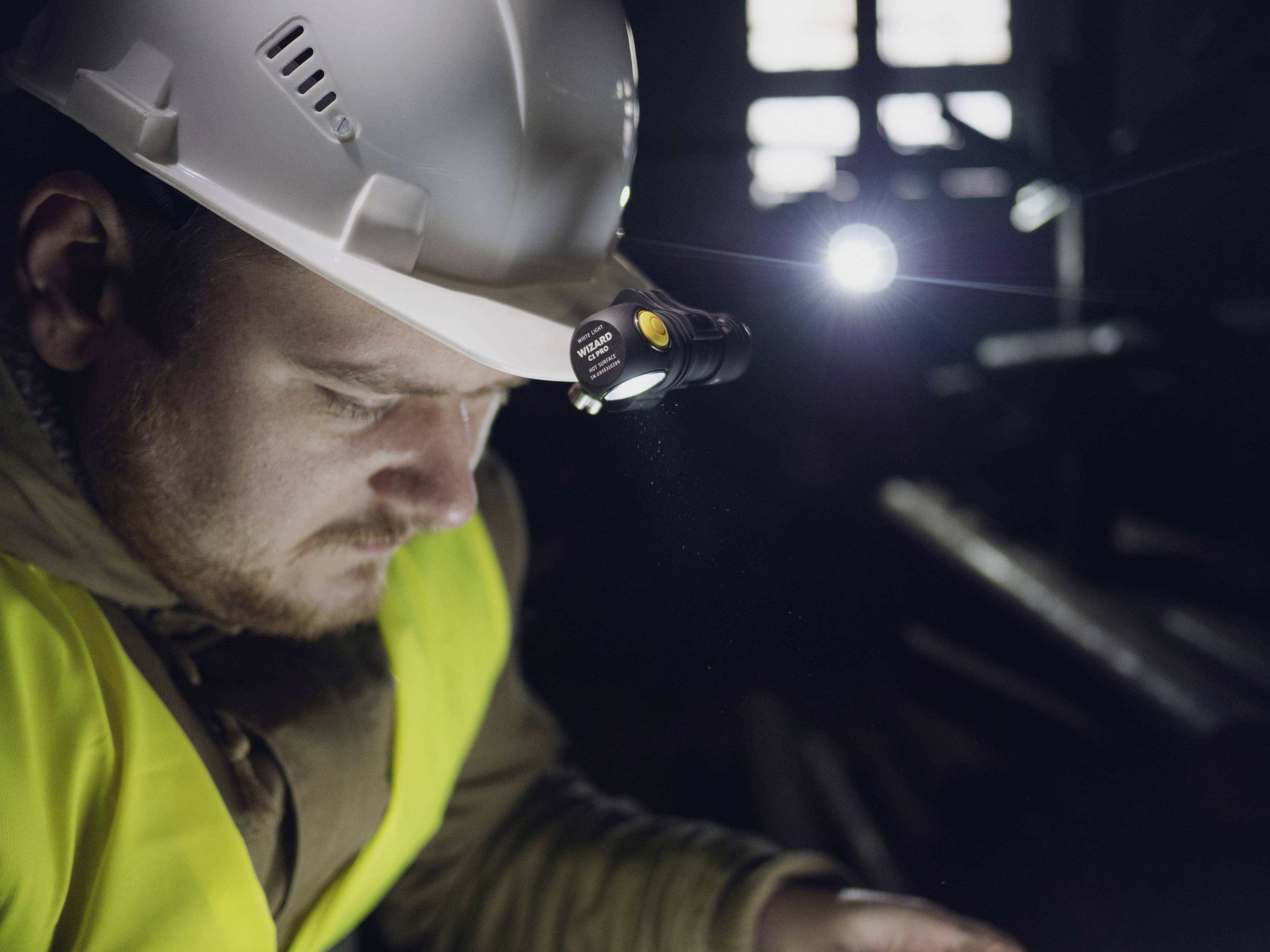A construction worker wearing a hard hat and head torch is working intently in a dark environment. He is wearing a high-visibility yellow vest.