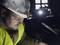 A construction worker wearing a hard hat and head torch is working intently in a dark environment. He is wearing a high-visibility yellow vest.