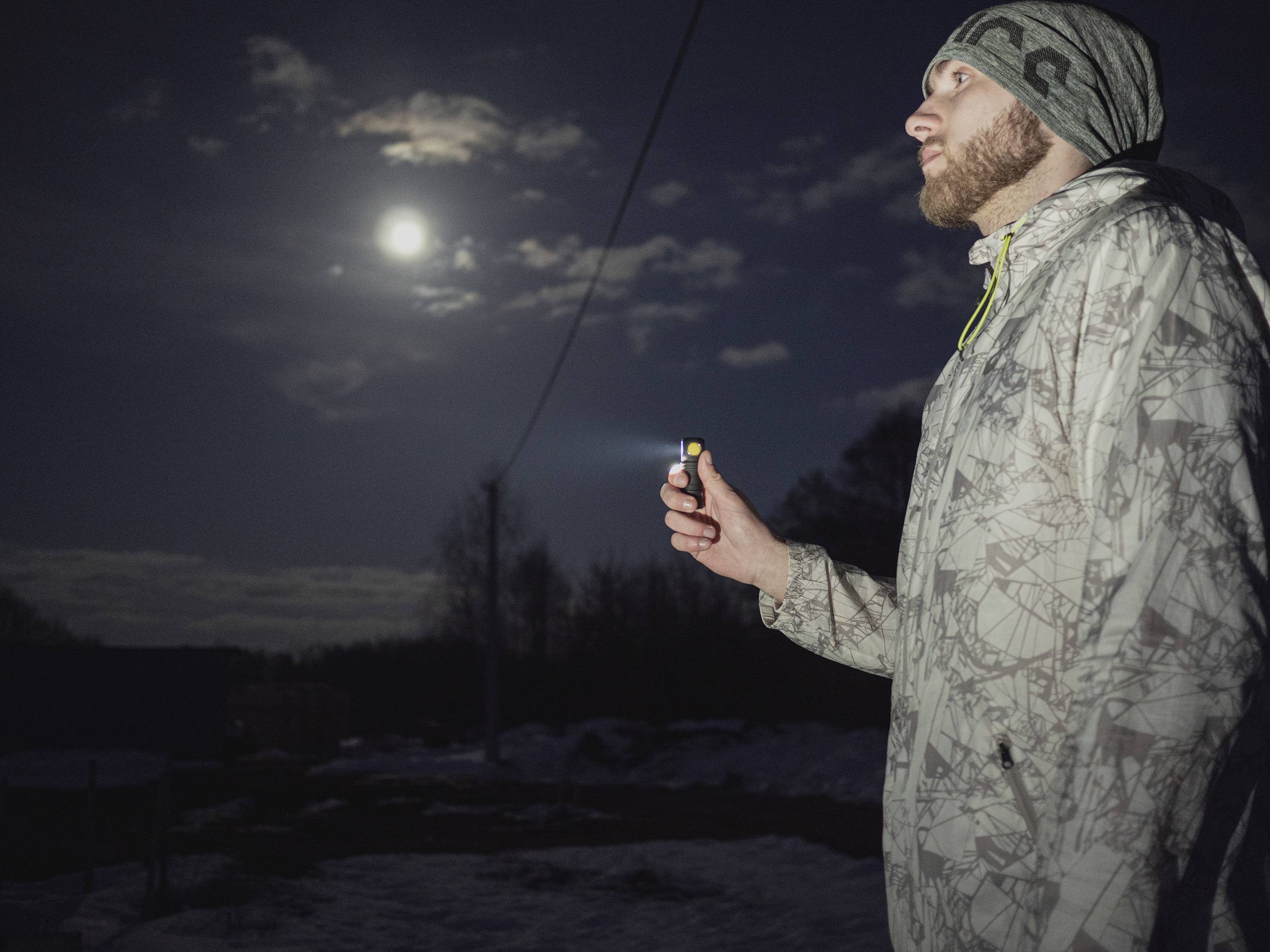 A man stands outside at night beneath a full moon, holding a small torch. It is wintry, with snow on the ground.