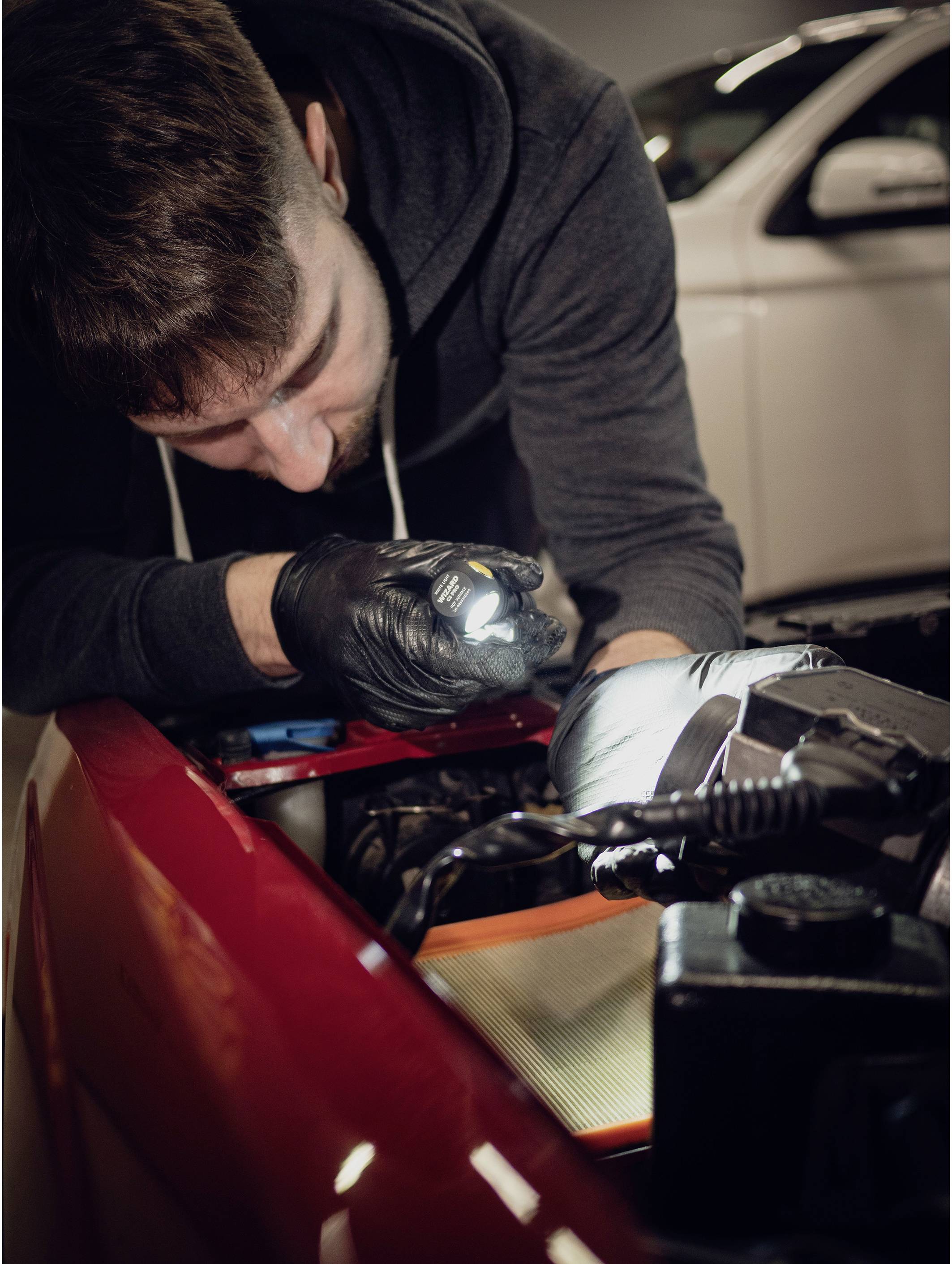 A mechanic repairs a car and shines a torch into the engine bay, focused on the work. A white car is in the background.