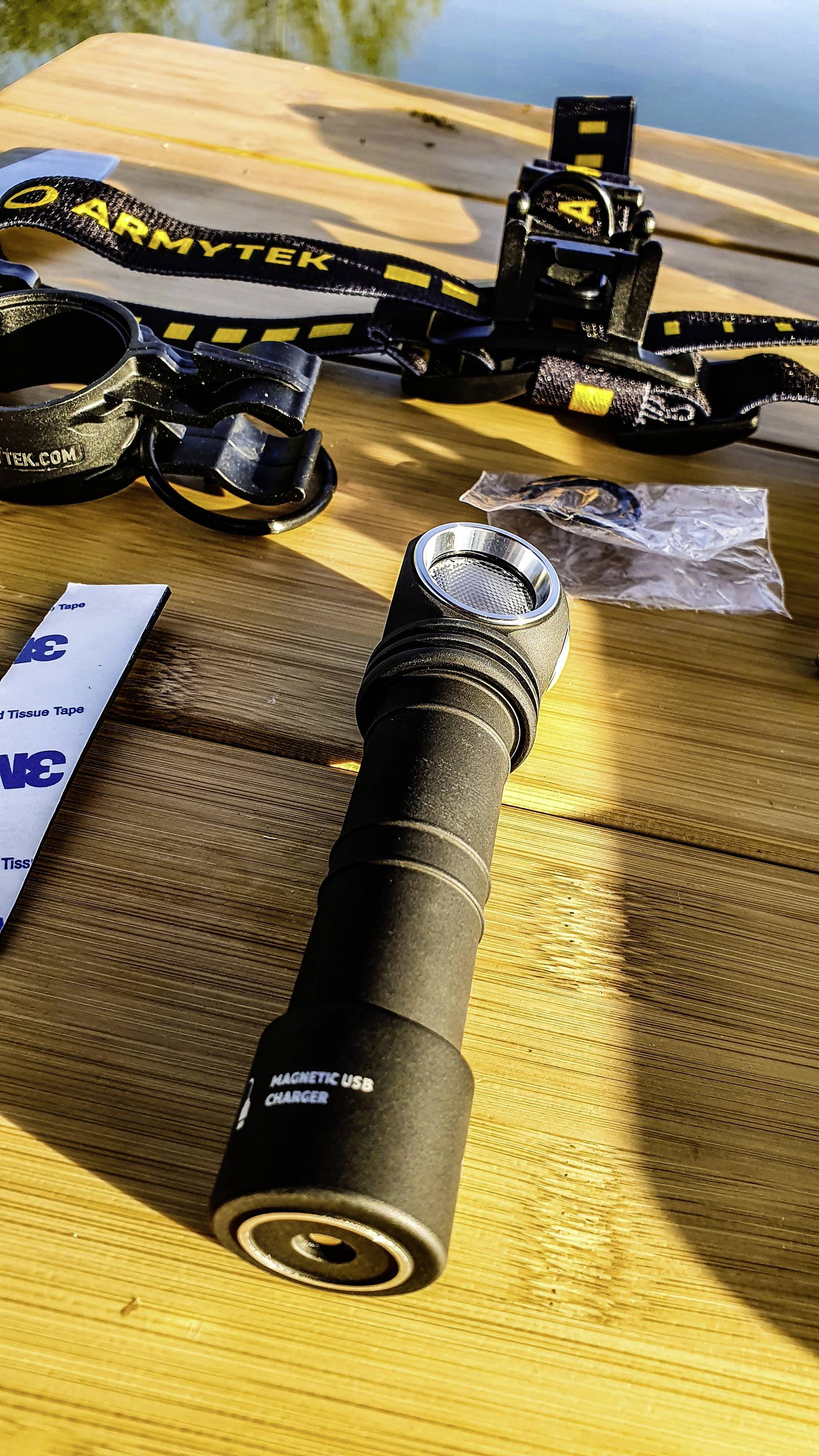 A torch lies on a wooden table next to a headband and accessories. A lake is reflected in the background.