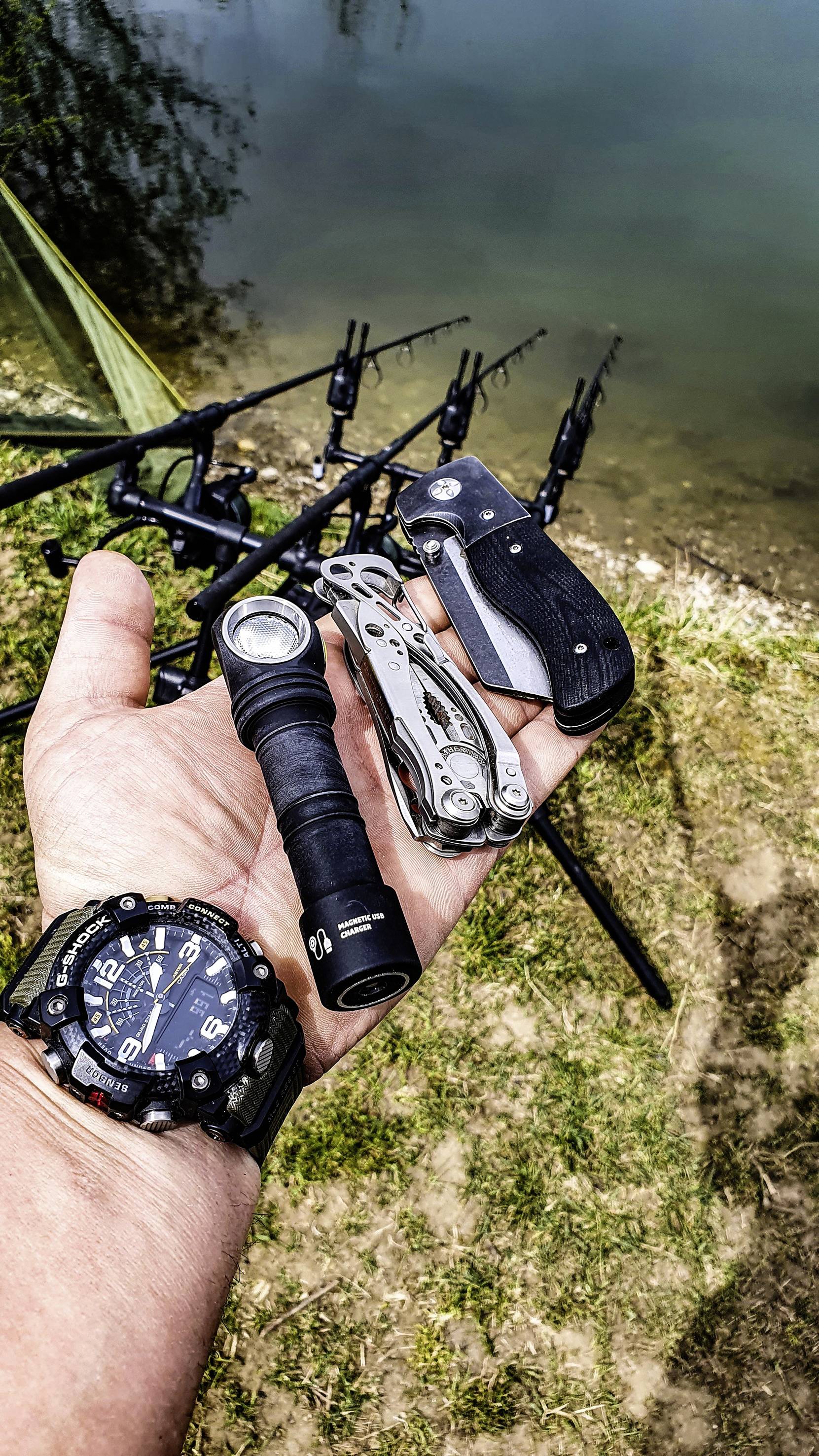 A hand holds a torch, a multi-tool and a penknife; fishing rods line the lakeshore in the background.