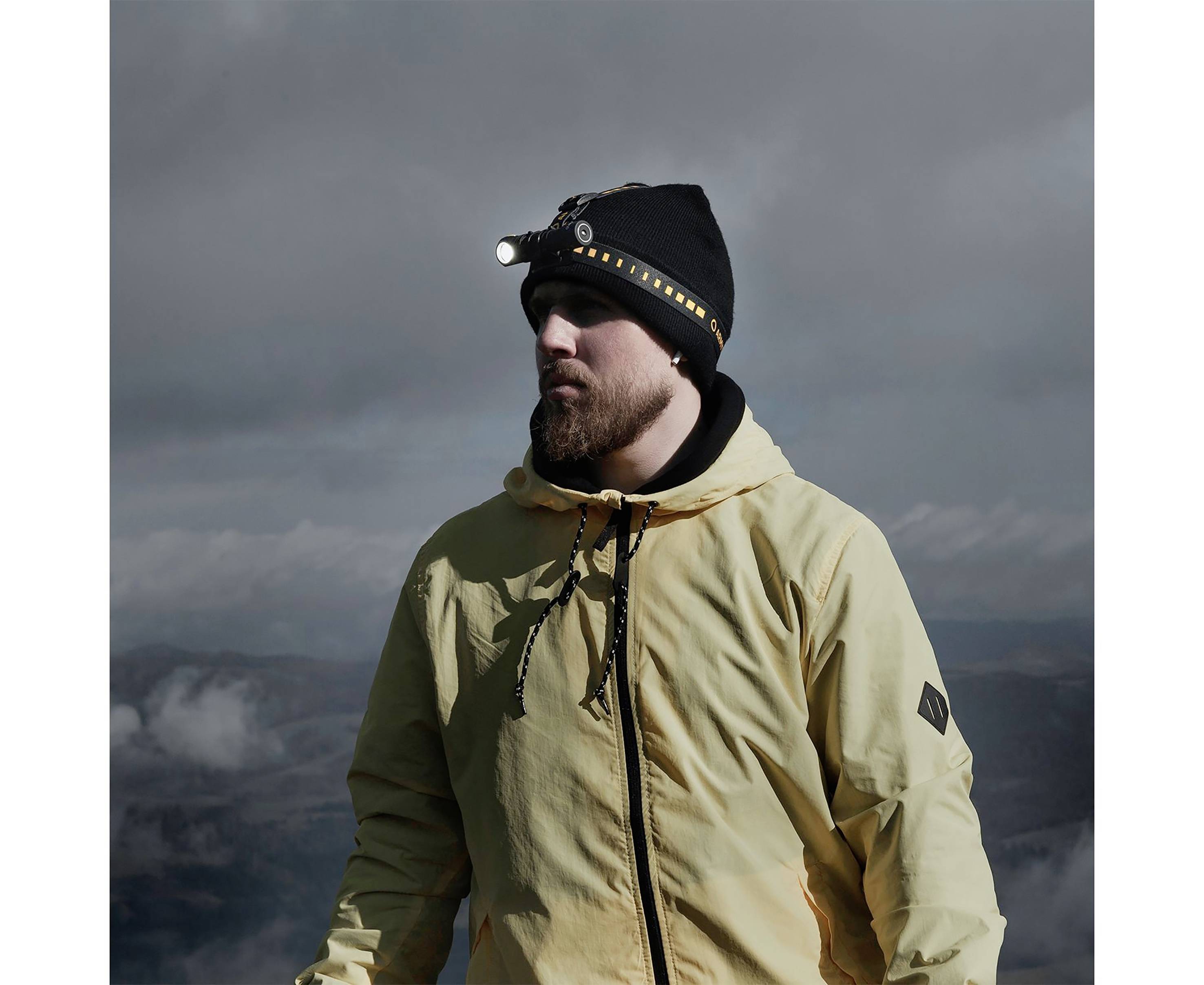 A person in a yellow jacket is standing outside. They are wearing a black beanie with a bright head torch, surrounded by cloudy skies.