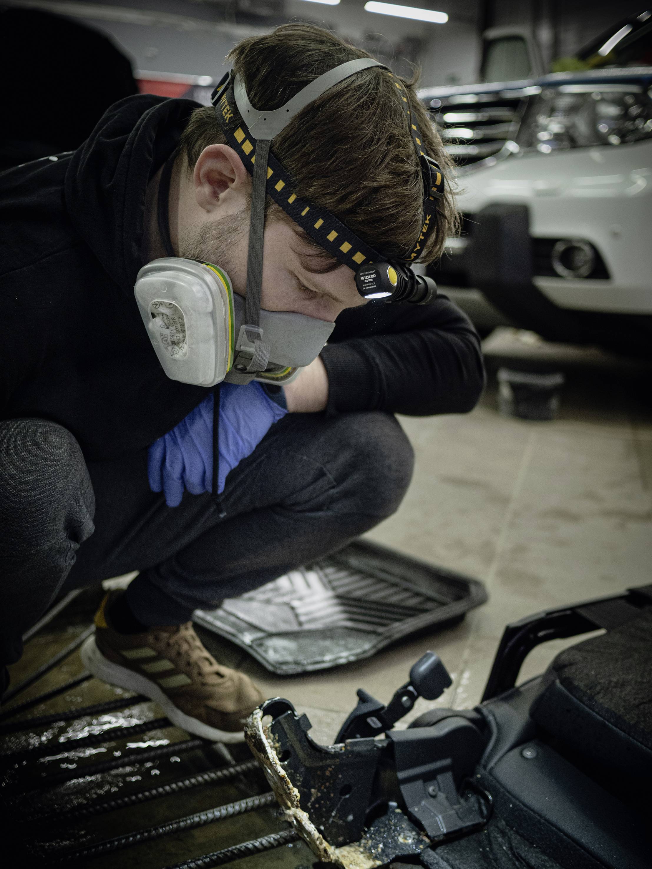 A man wearing a protective mask and a head torch sits checking an automotive part in a workshop. Cars are parked in the background.
