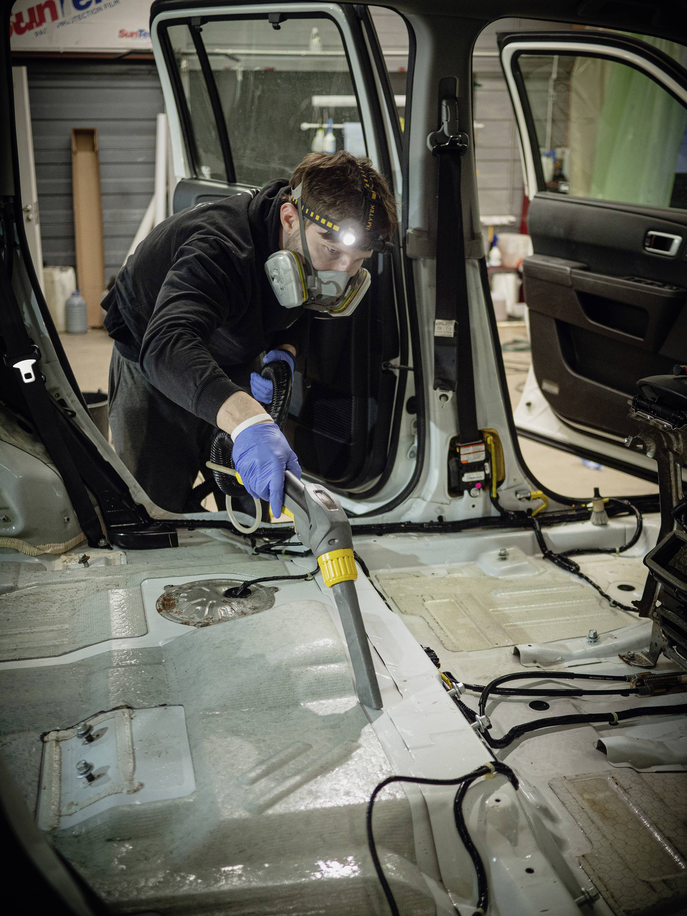 A person using a vacuum cleaner inside a car with no vehicle, wearing a protective mask and gloves.