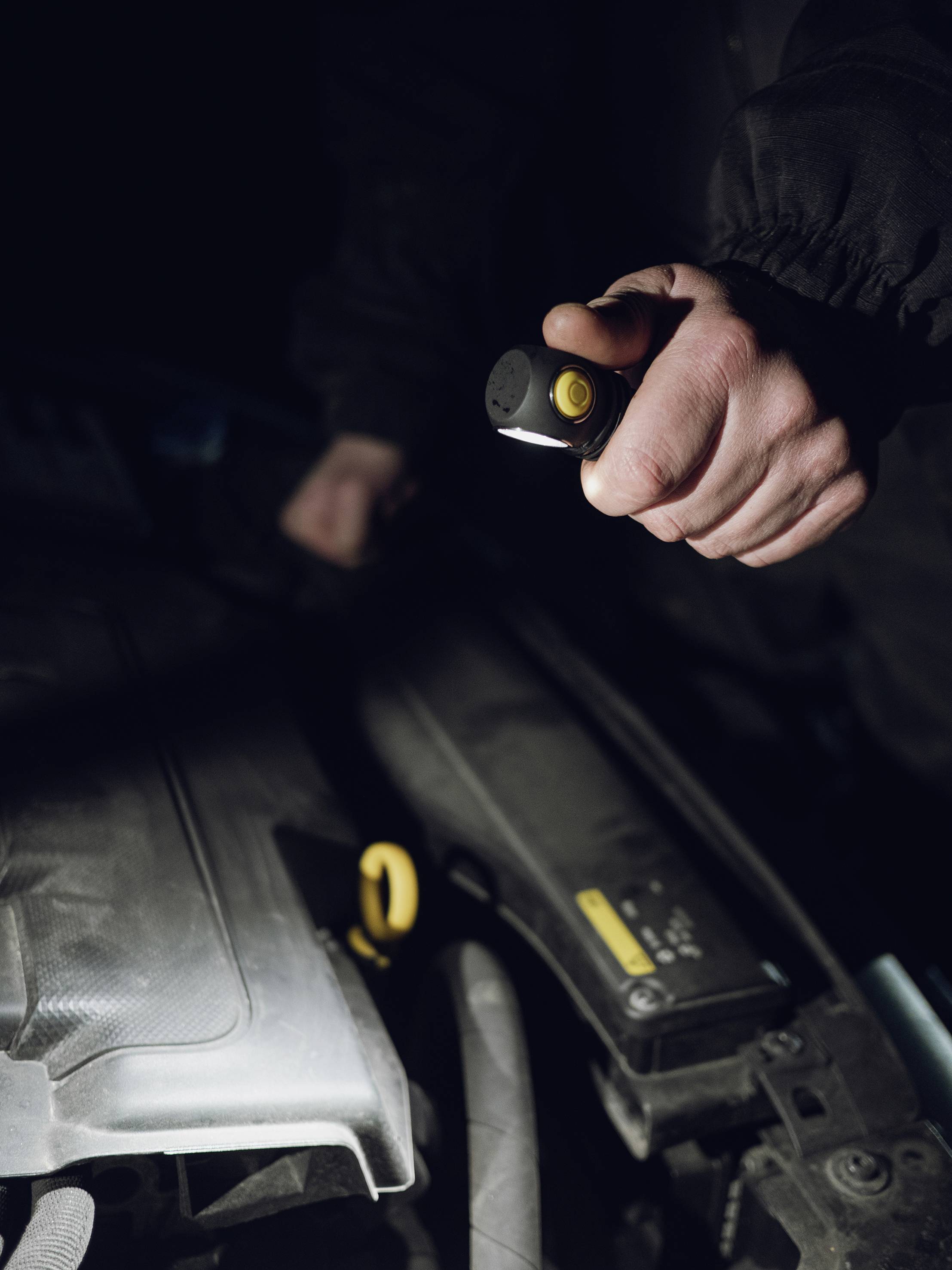 A mechanic is examining the engine bay of a car with a torch in low light conditions.