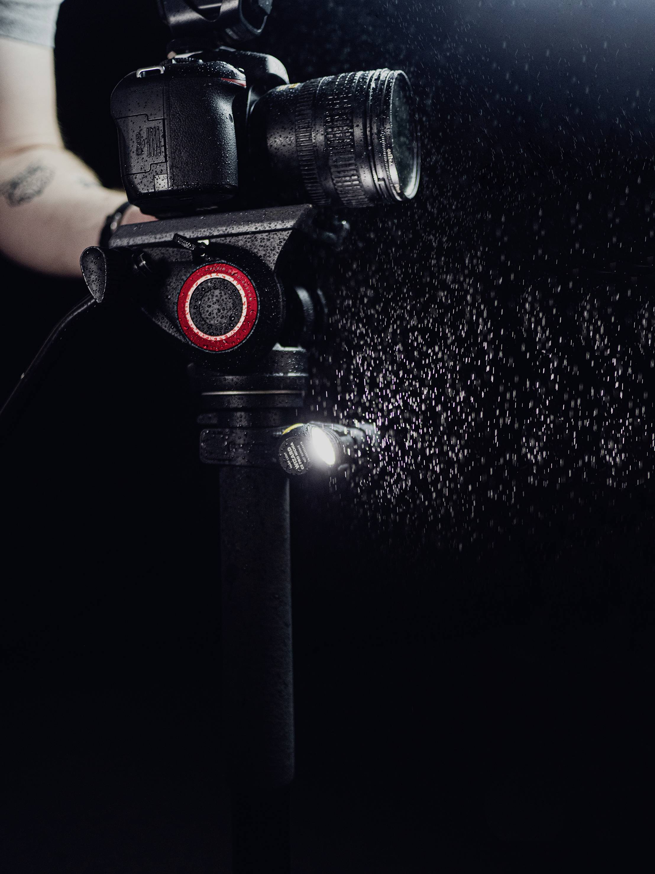 Camera on a tripod, photographed from an angled overhead view. A hand adjusts the settings, with water droplets visible in the background.