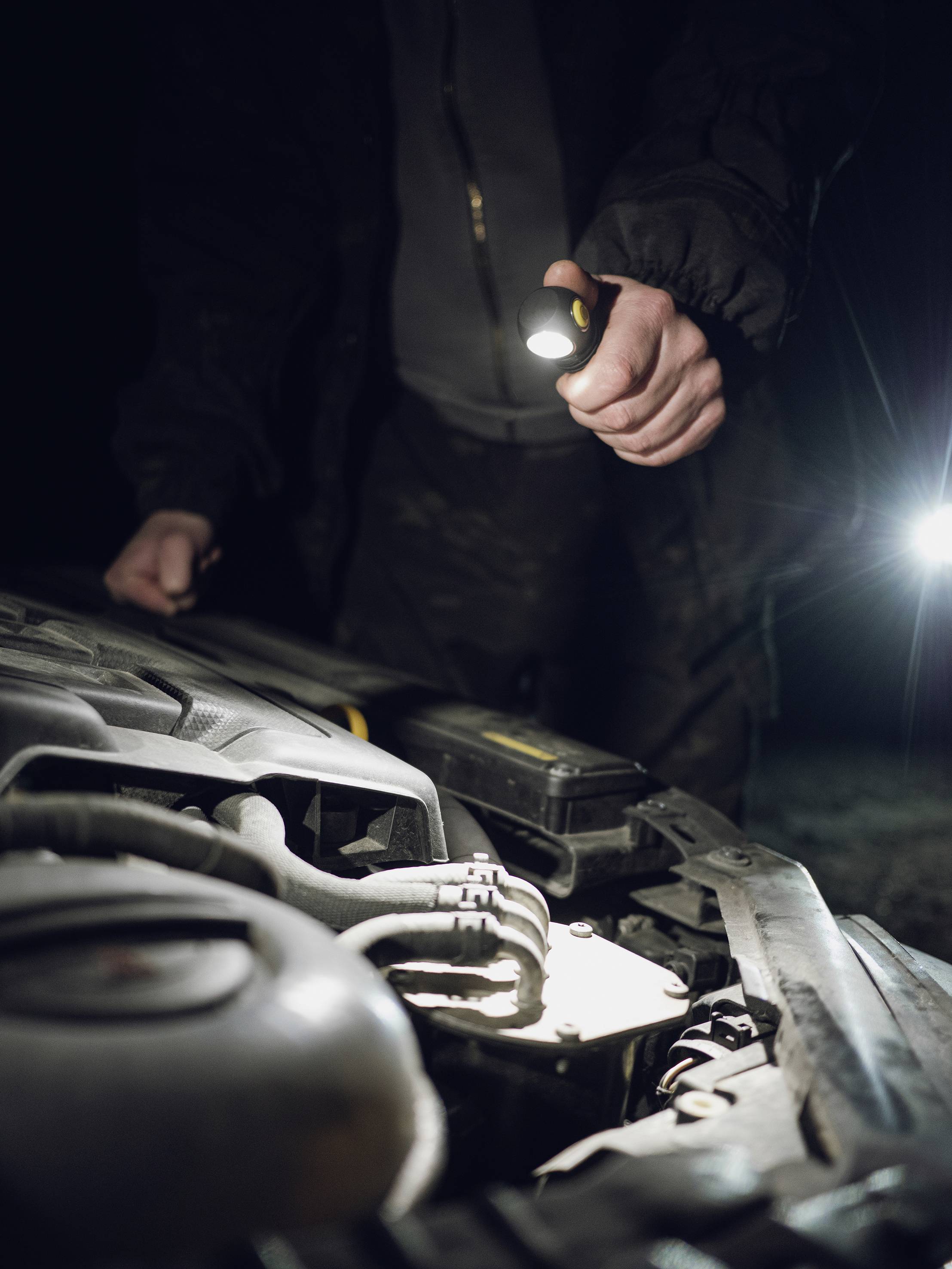 "A person is examining the engine compartment of a car at night using a torch."<br><br>Note: In British English, "flashlight" would be replaced with "torch", which is the standard term used in the UK.