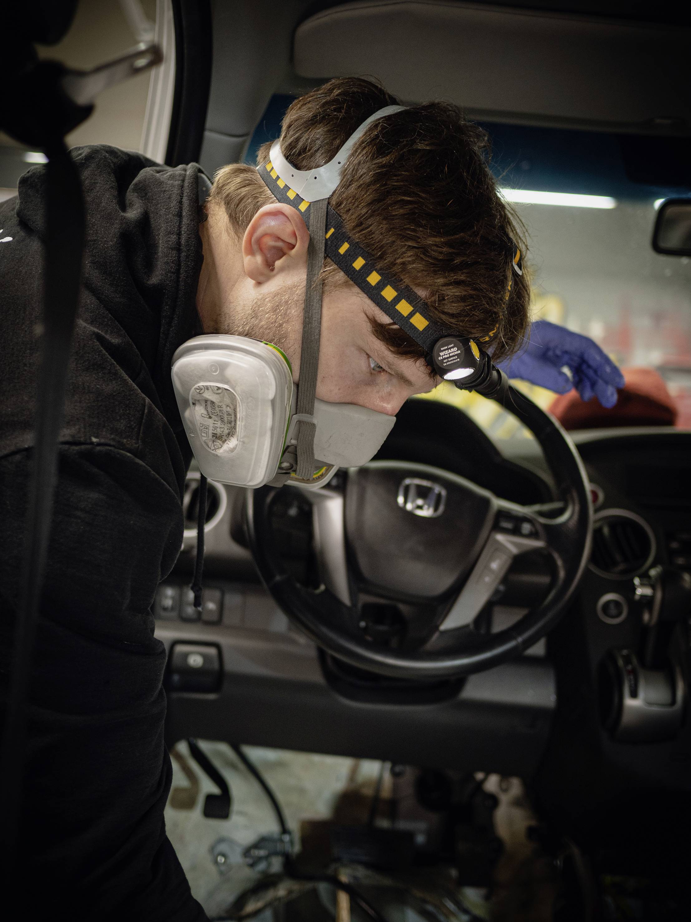 A man wearing a respiratory mask and head torch is working intently inside a car, presumably addressing a technical issue.