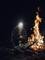 A person wearing a head torch kneels beside a large campfire in the woods at night. They are adding branches to the fire.