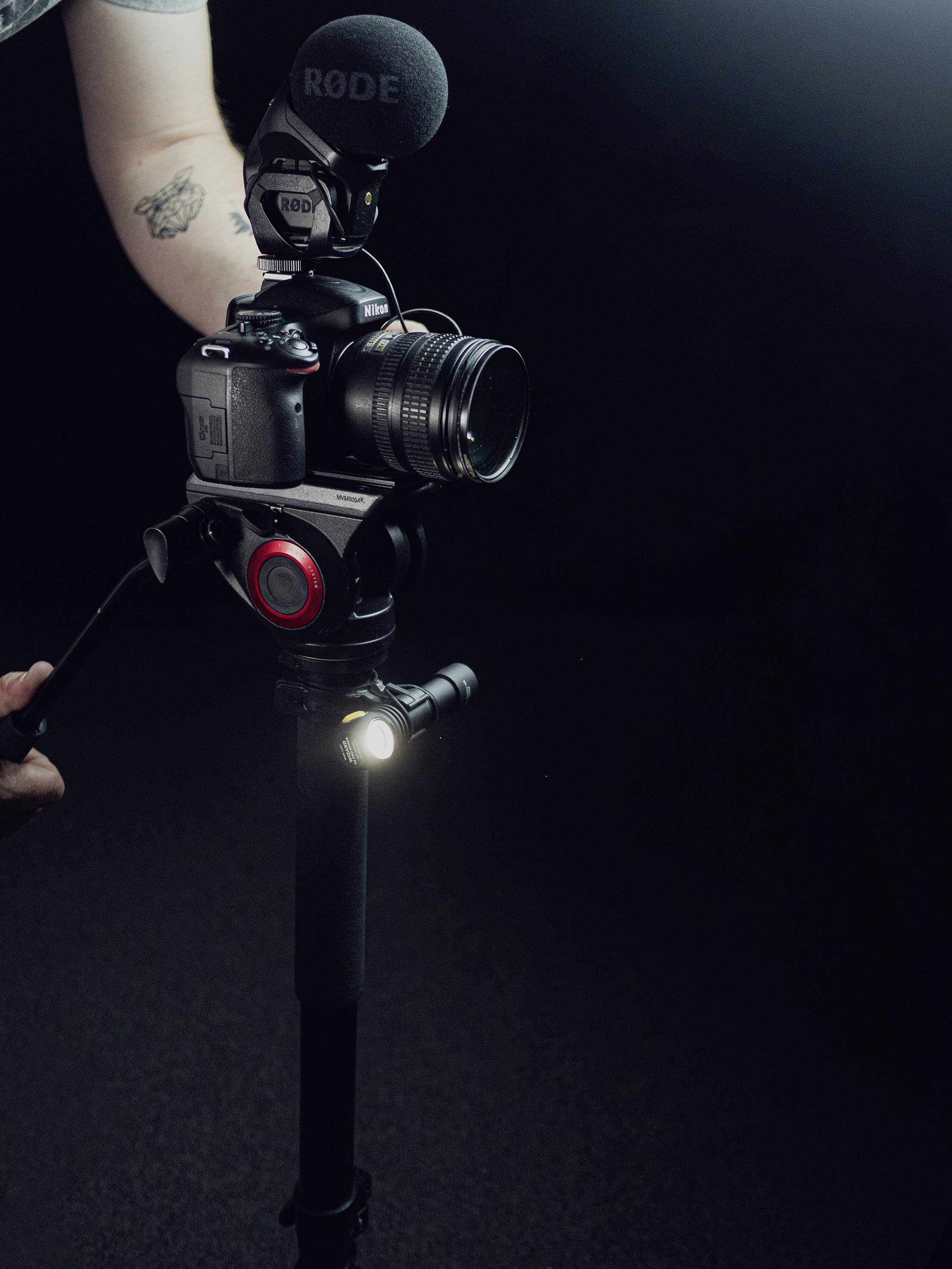 A camera with a microphone mounted on a tripod stands in a dark room. A person is adjusting the tripod.