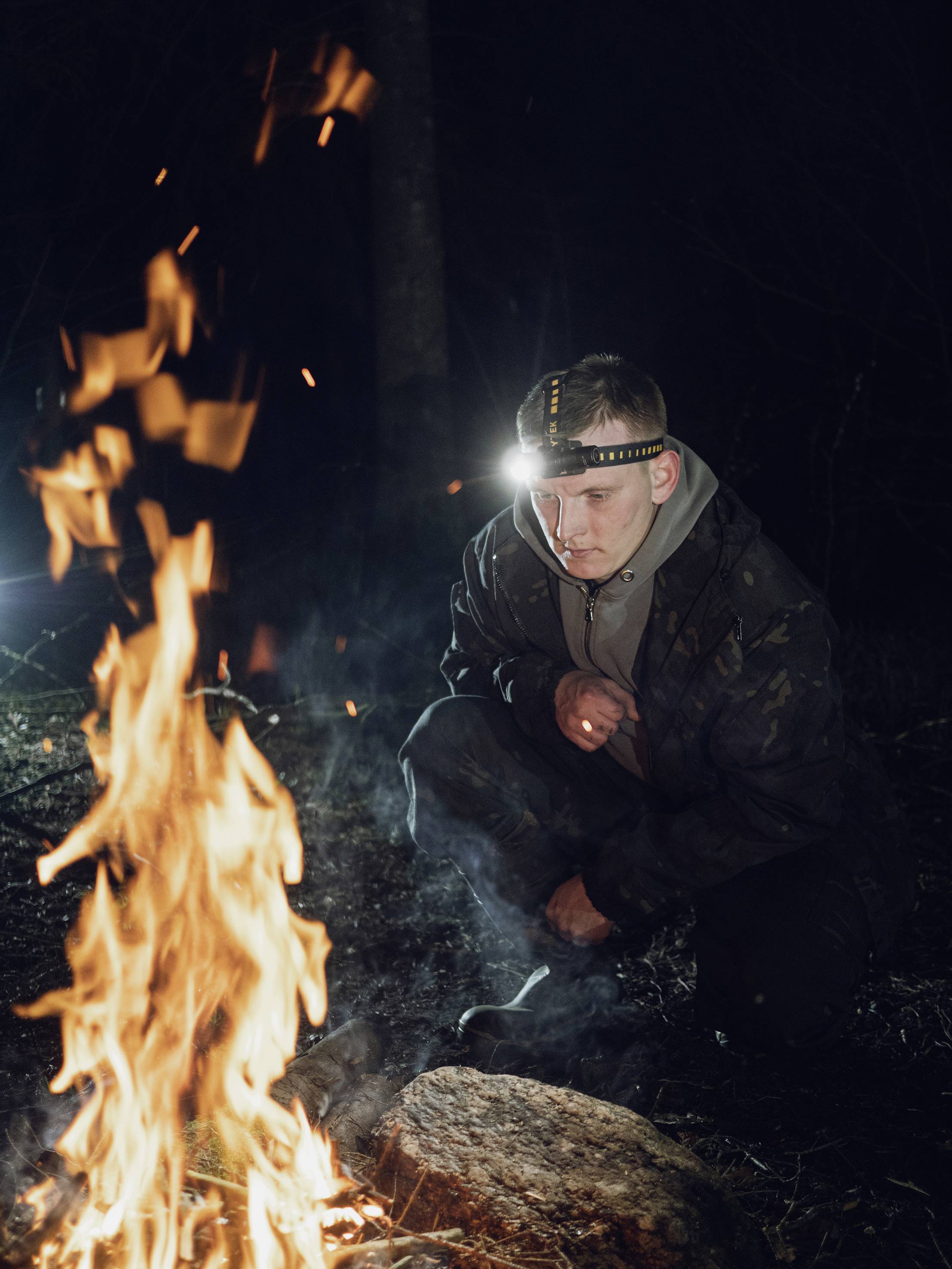 A person with a head torch crouches by the campfire at night in the woods, surrounded by trees.