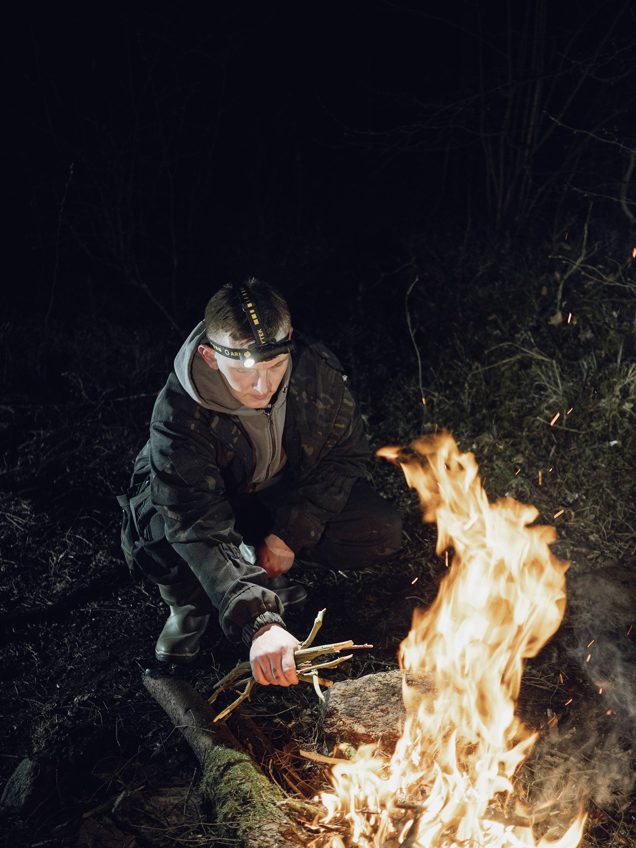 A person in outdoor clothing lights a campfire at night using a fire starter.