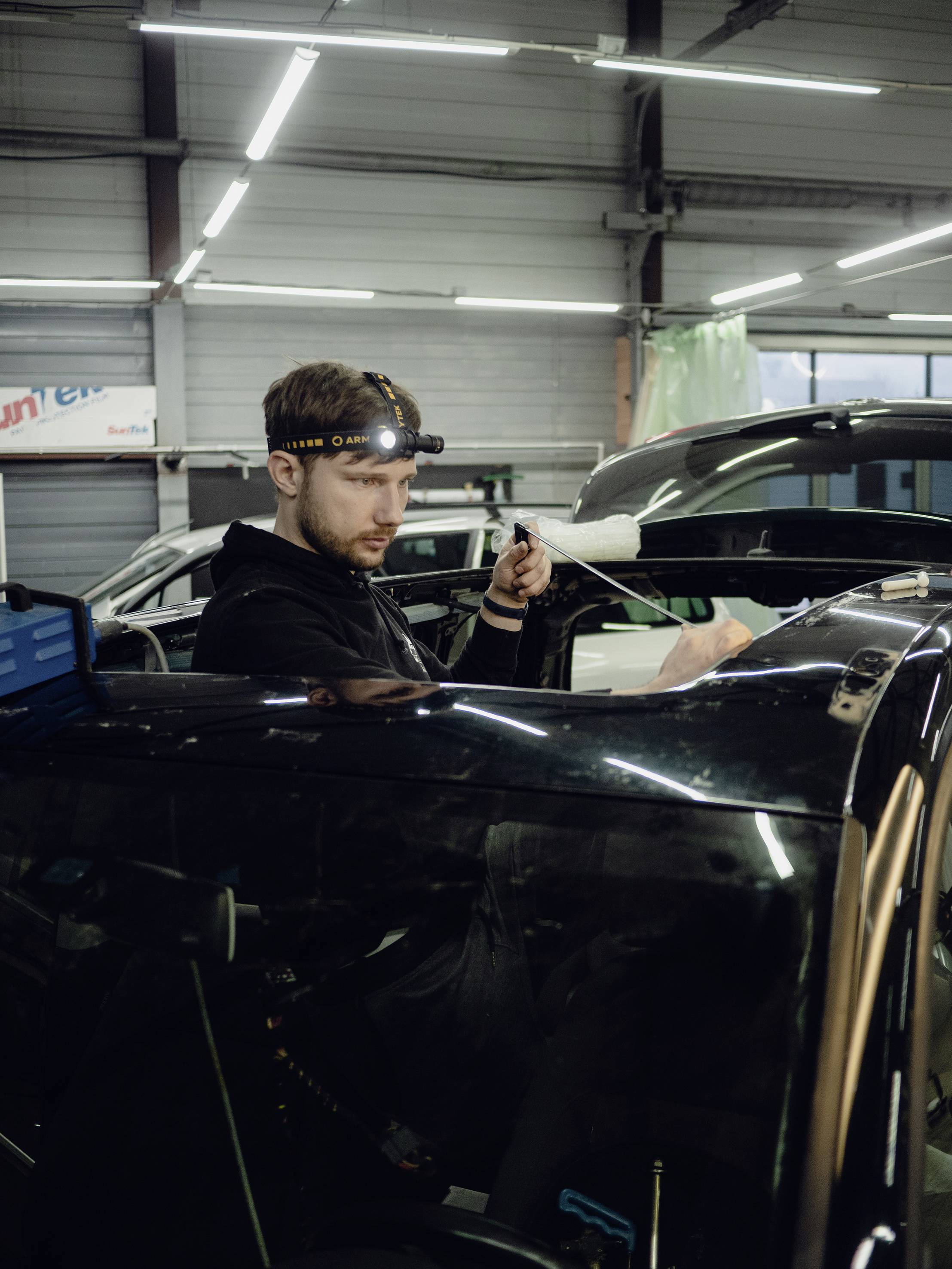 A man is working in a car garage. He is wearing a head torch and repairing a black car in a workshop environment.