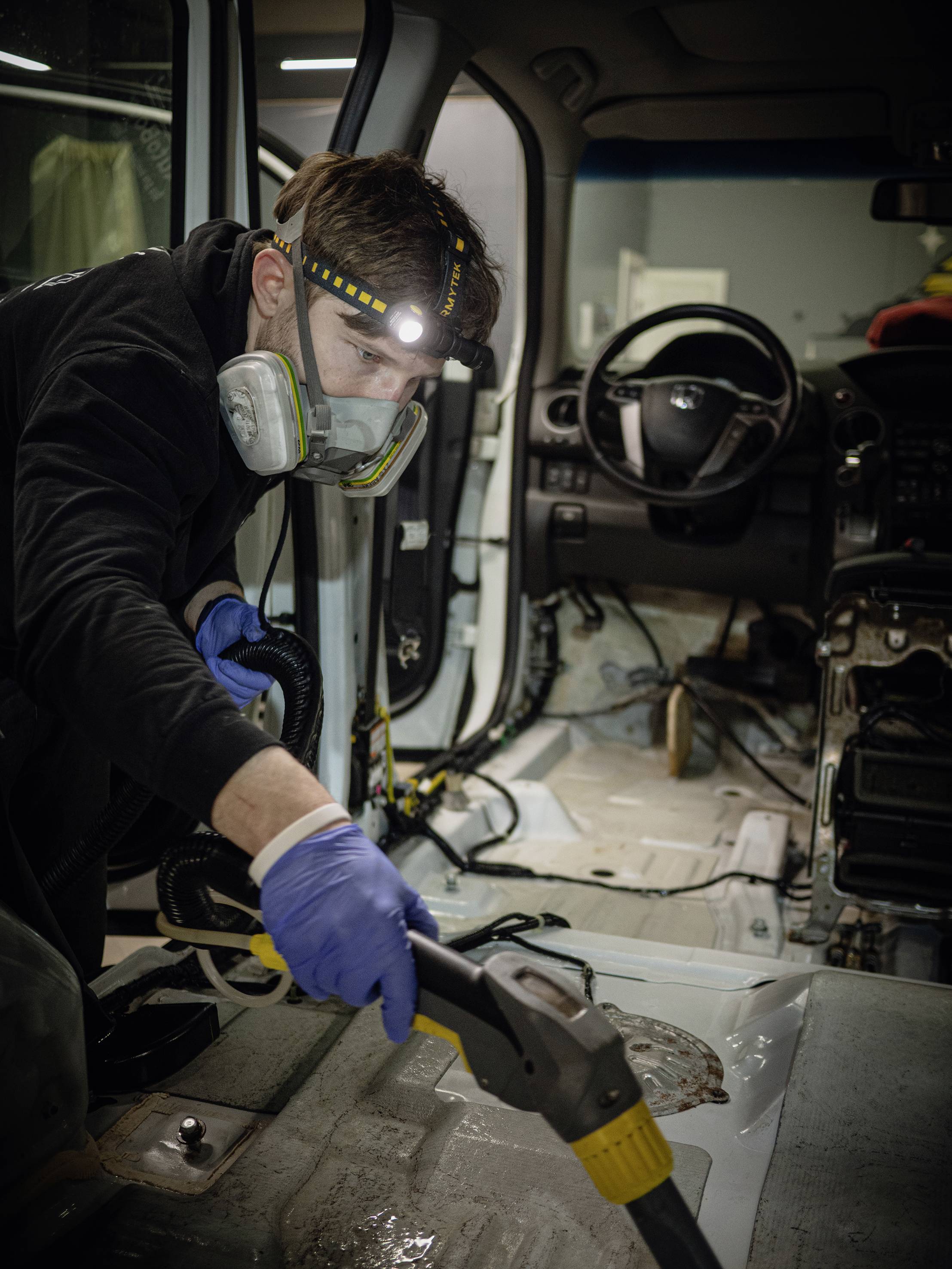 A person wearing protective clothing is cleaning the interior of a car with a steam cleaner. The interior is cleared out.