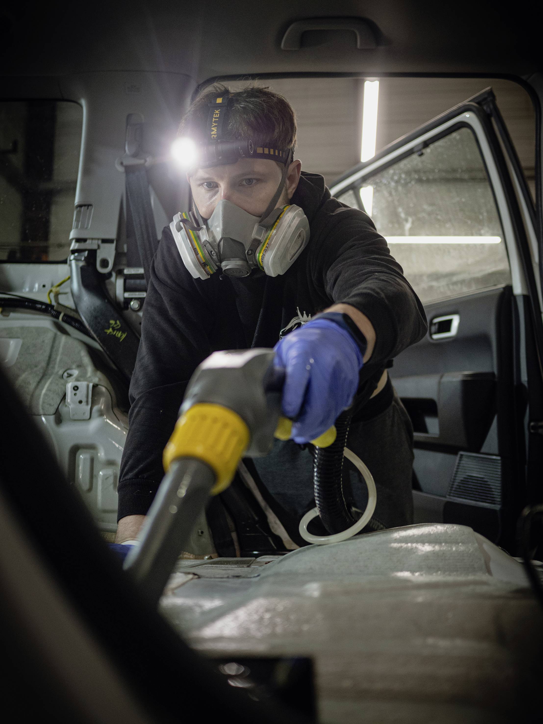 A man is cleaning the interior of a car with a machine. He is wearing a respirator, gloves, and a head torch to facilitate the work.