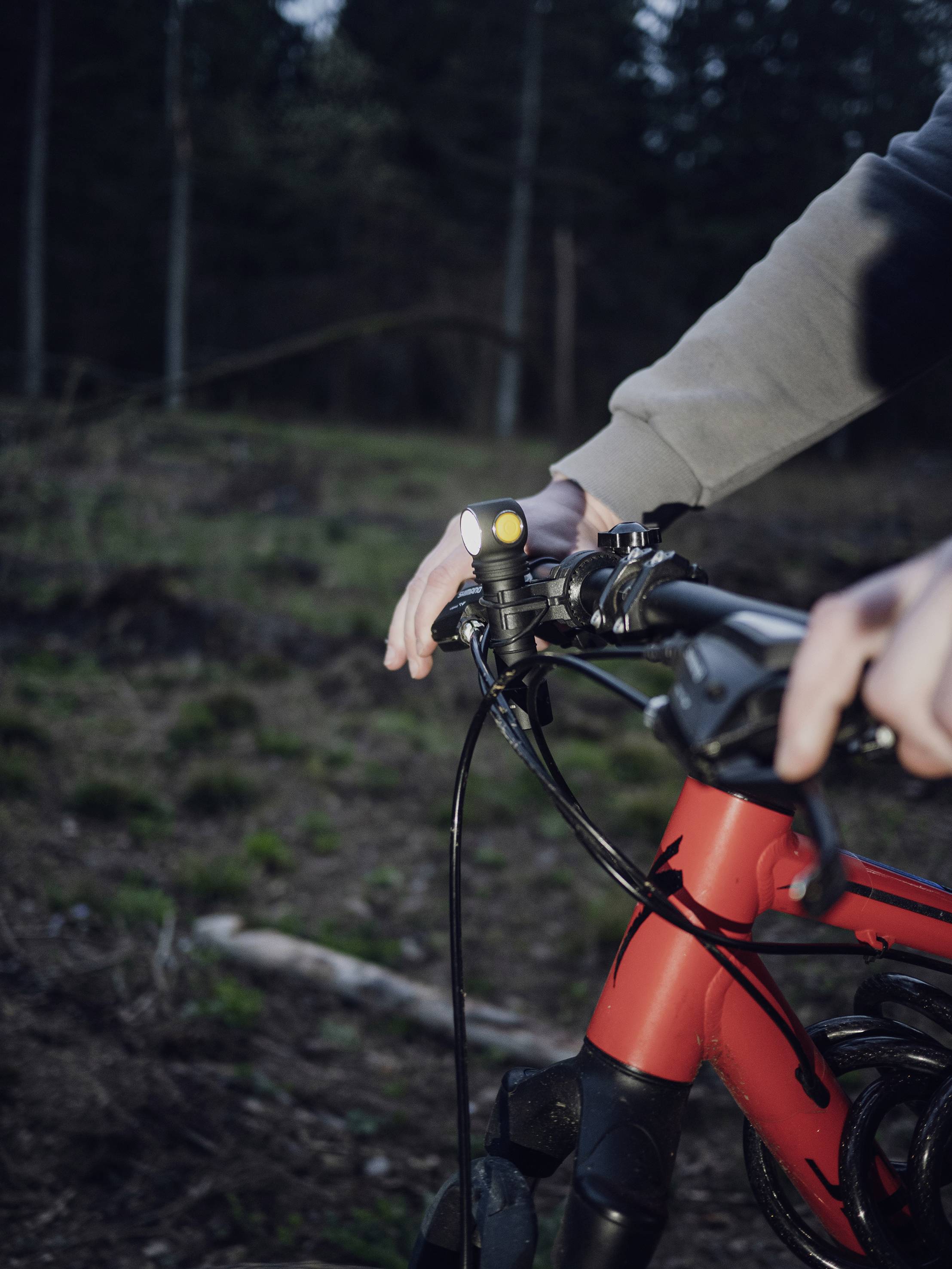 A person is holding a red bicycle in the woods. The focus is on the hand on the handlebar and a small light. Trees can be seen in the background.