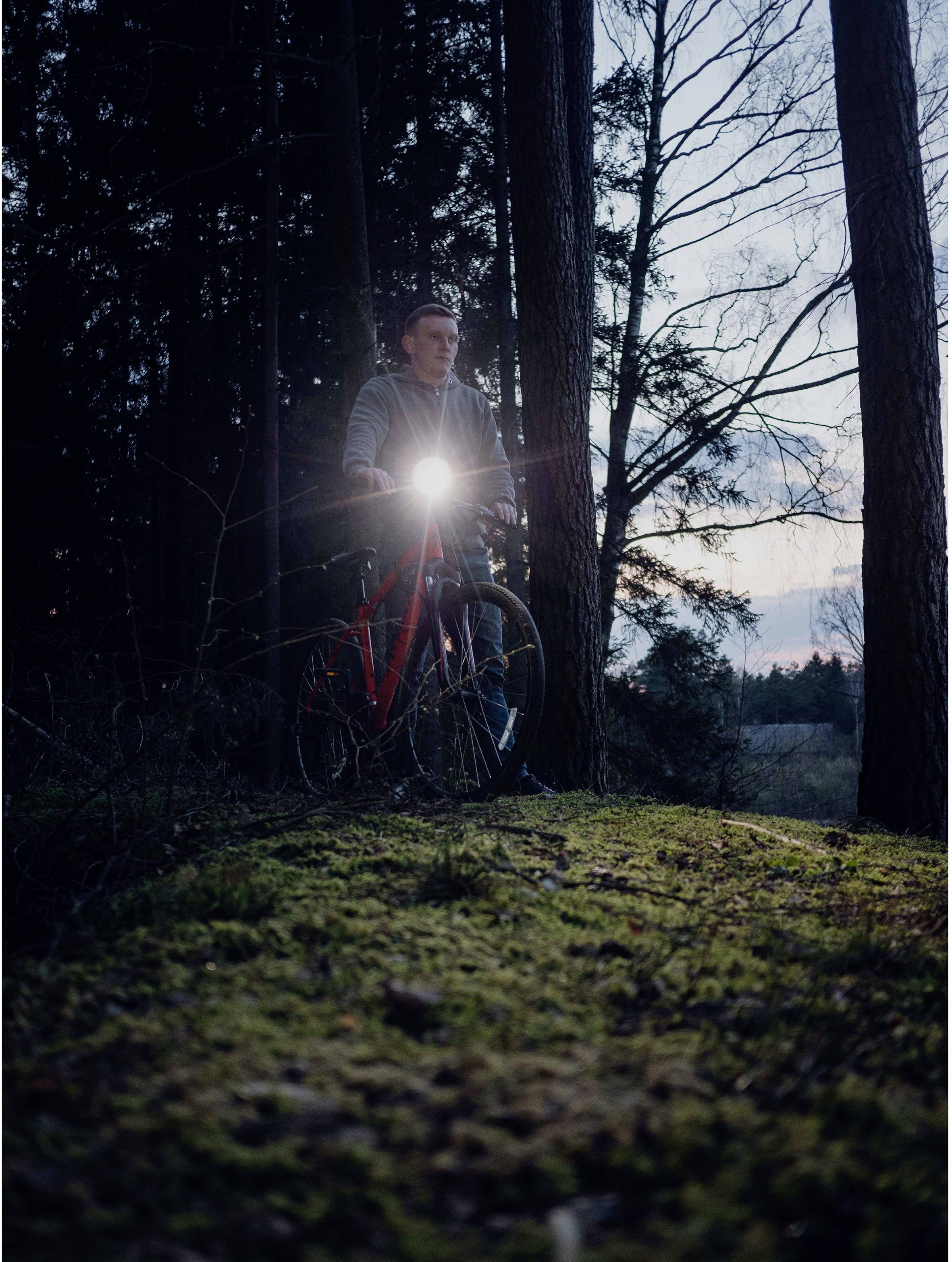 A person is standing with a bicycle in the woods, the bicycle's front light switched on. It is dusk.