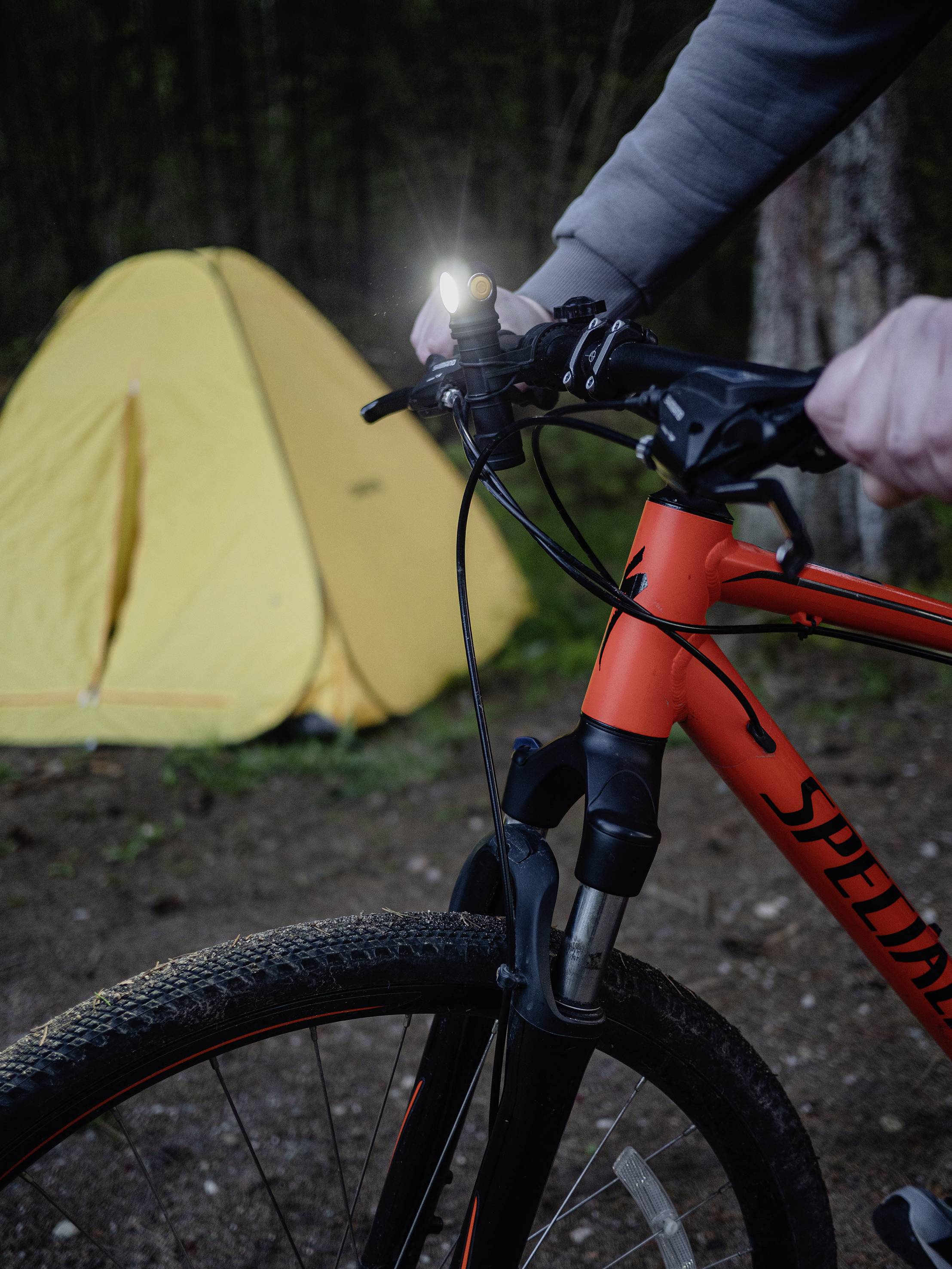 A person is holding a bicycle handlebar with a light switched on. A yellow tent stands in the background. The scene is set outdoors.