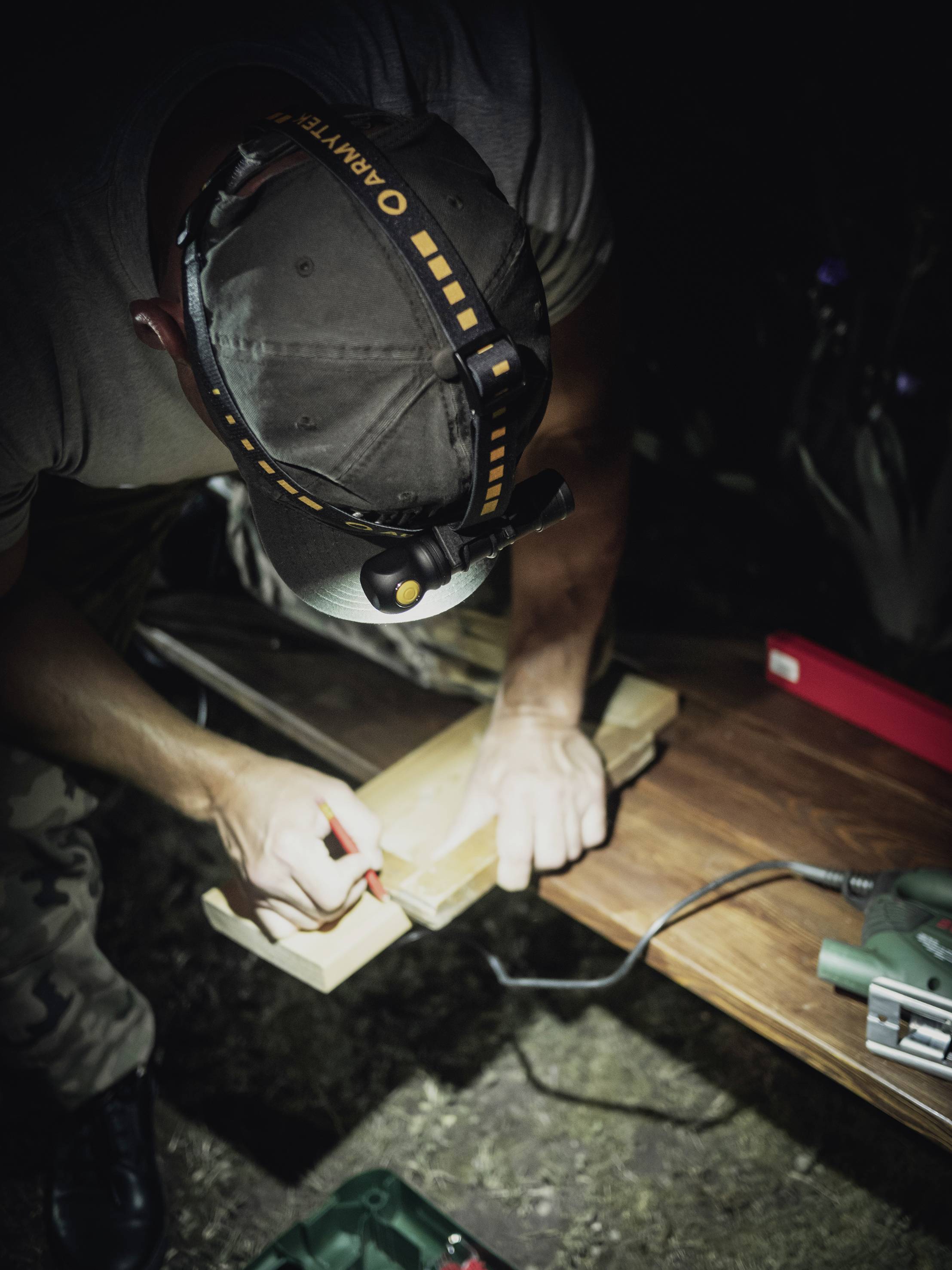 A person in a dark environment draws a line on a piece of wood, illuminated by a head torch. Tools are lying nearby.