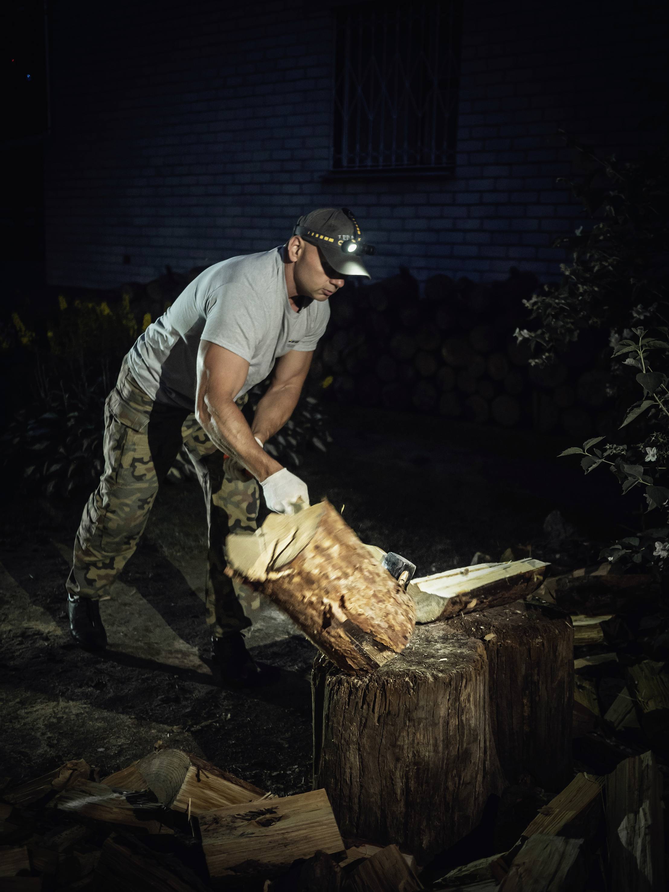 A man in camouflage trousers is chopping wood on a tree stump at night. He is wearing a head torch and gloves. In the background, a woodpile is visible.