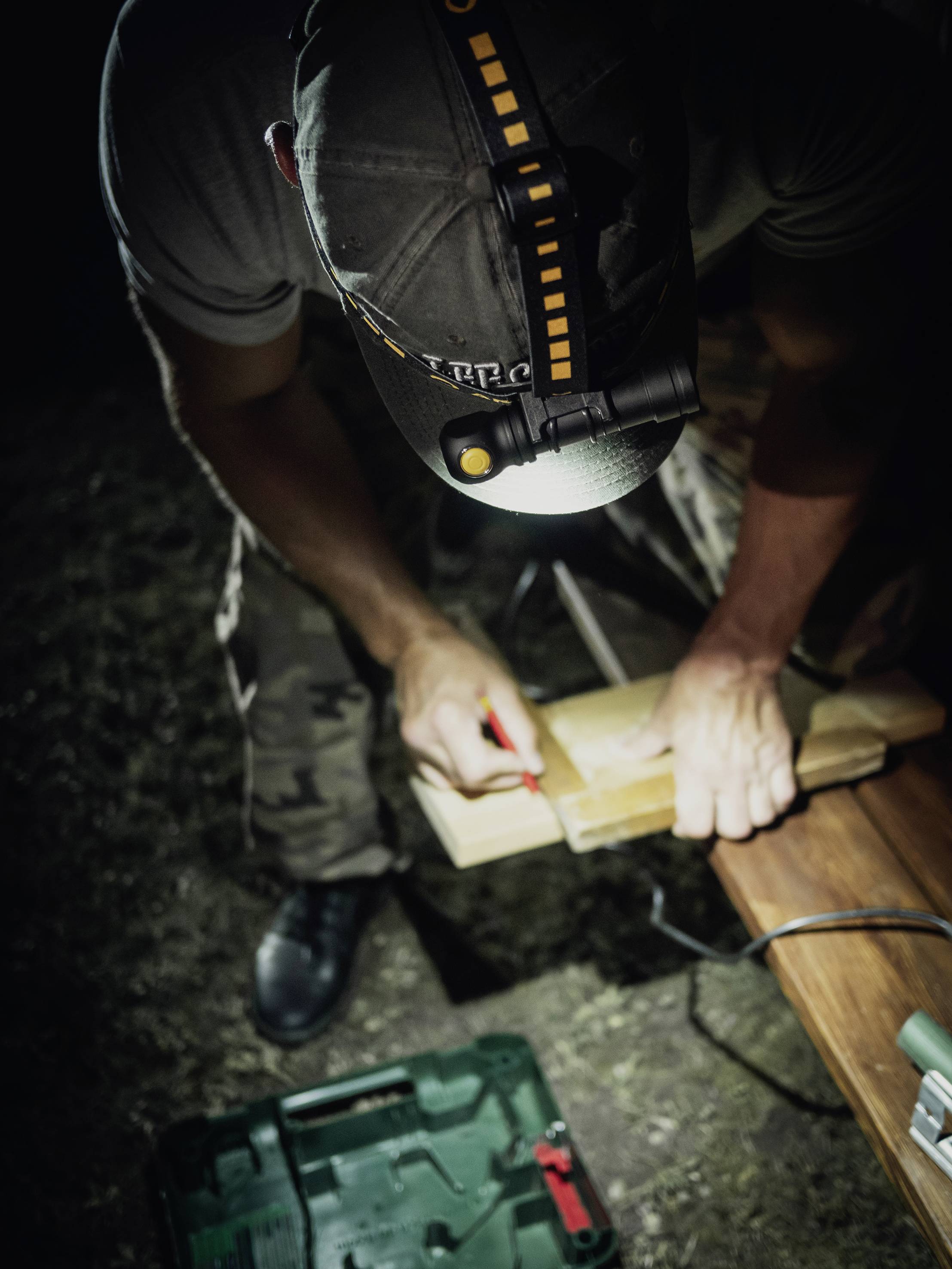 A person is working outdoors at night with a head torch on a wooden board. An open toolbox lies on the grass in front of them.