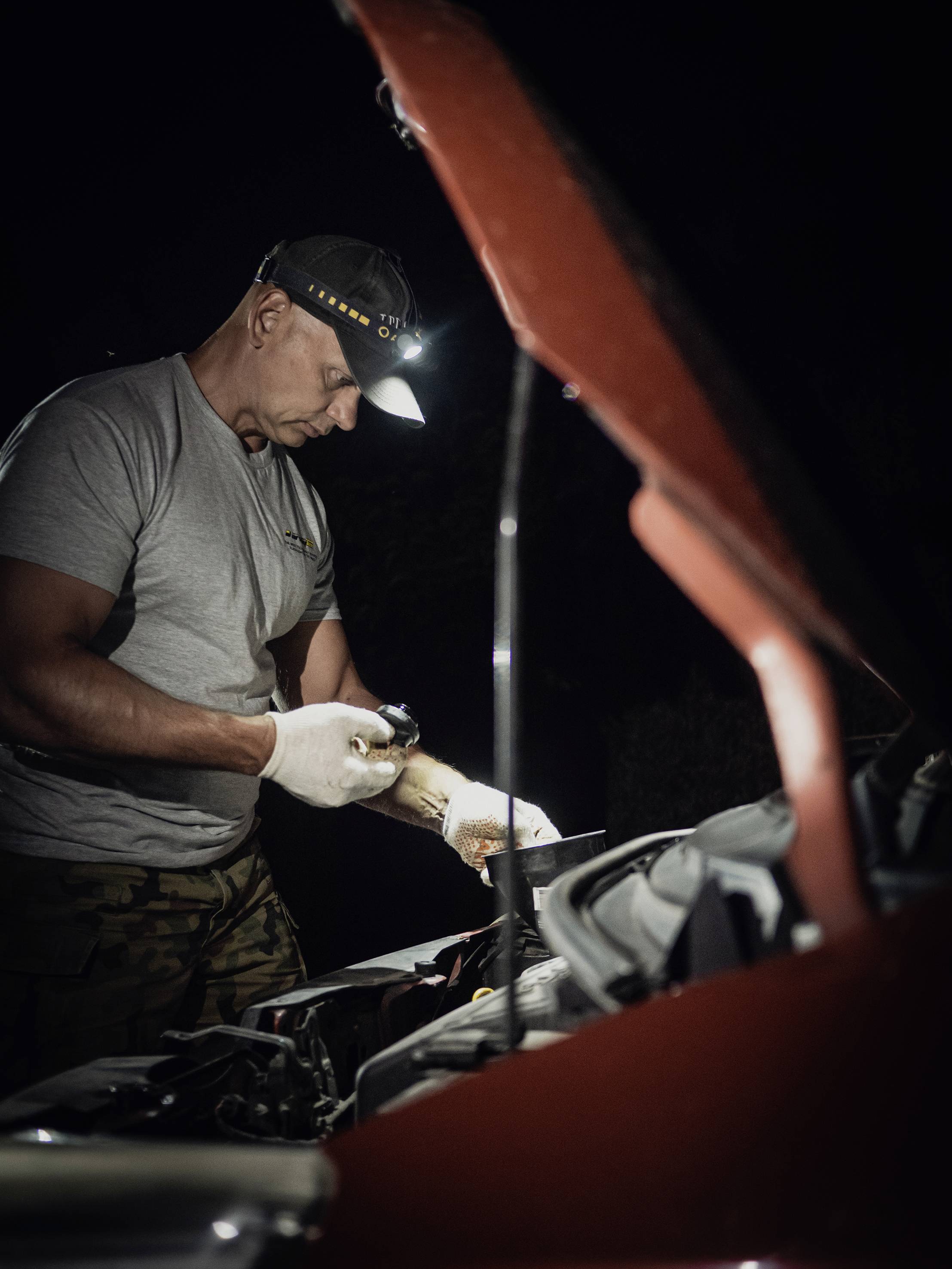 A man in a grey t-shirt is repairing a car's engine at night with a head torch, with the bonnet open.