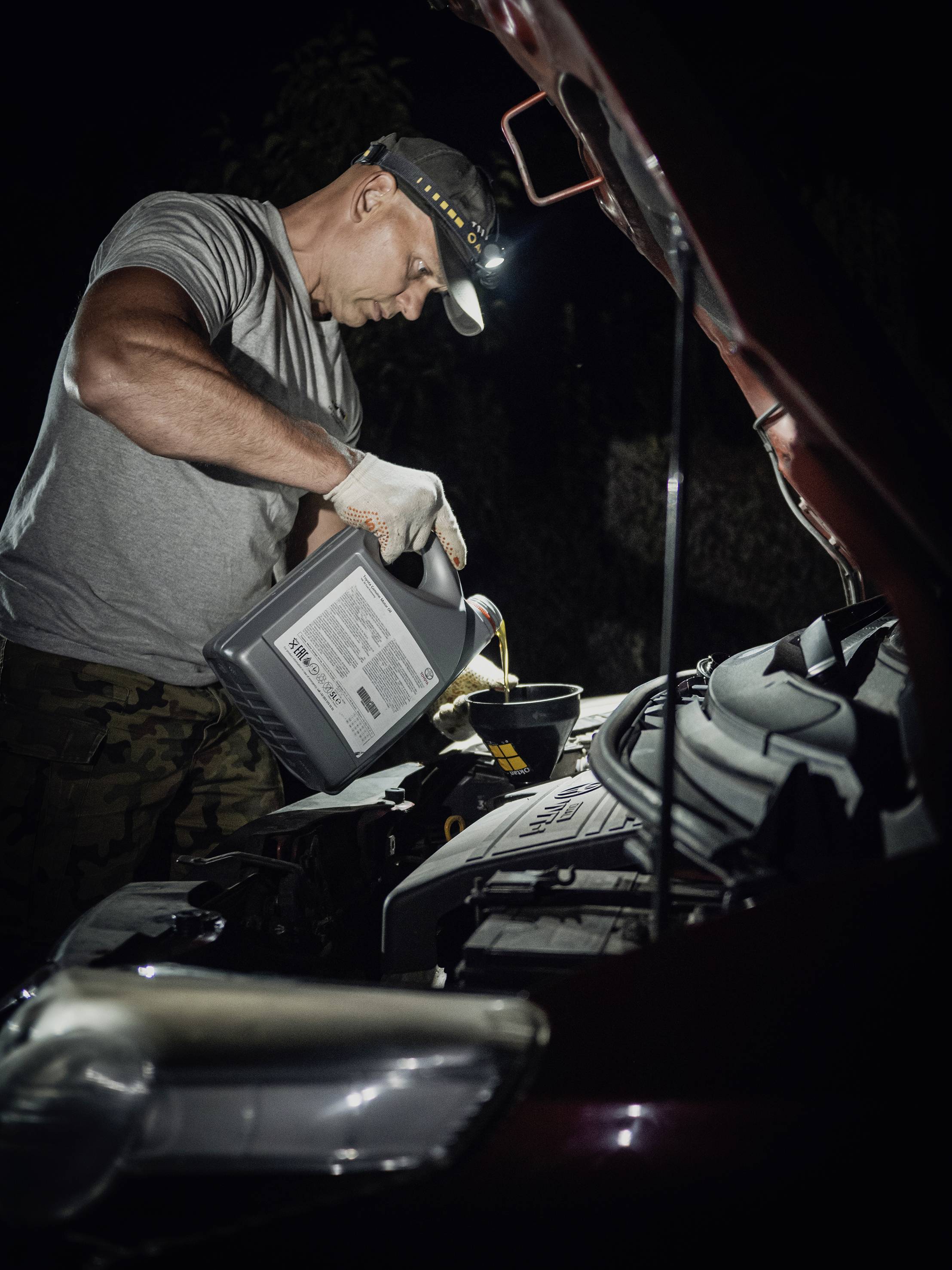 A man in workwear is filling oil into a car's engine in the dark. He is wearing a head torch and working with concentration.