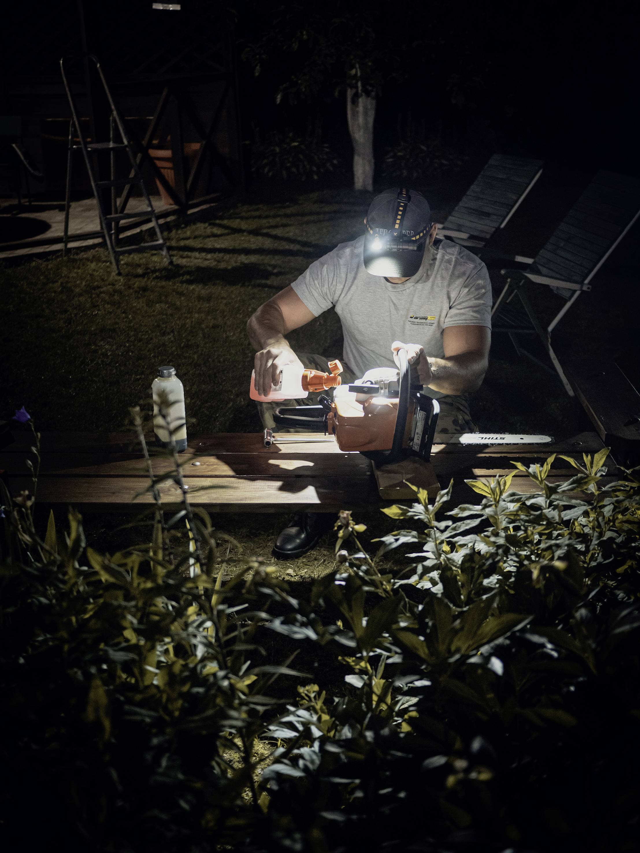 A person wearing a head torch is working outside at night at a table, surrounded by plants. They are focused on a project.