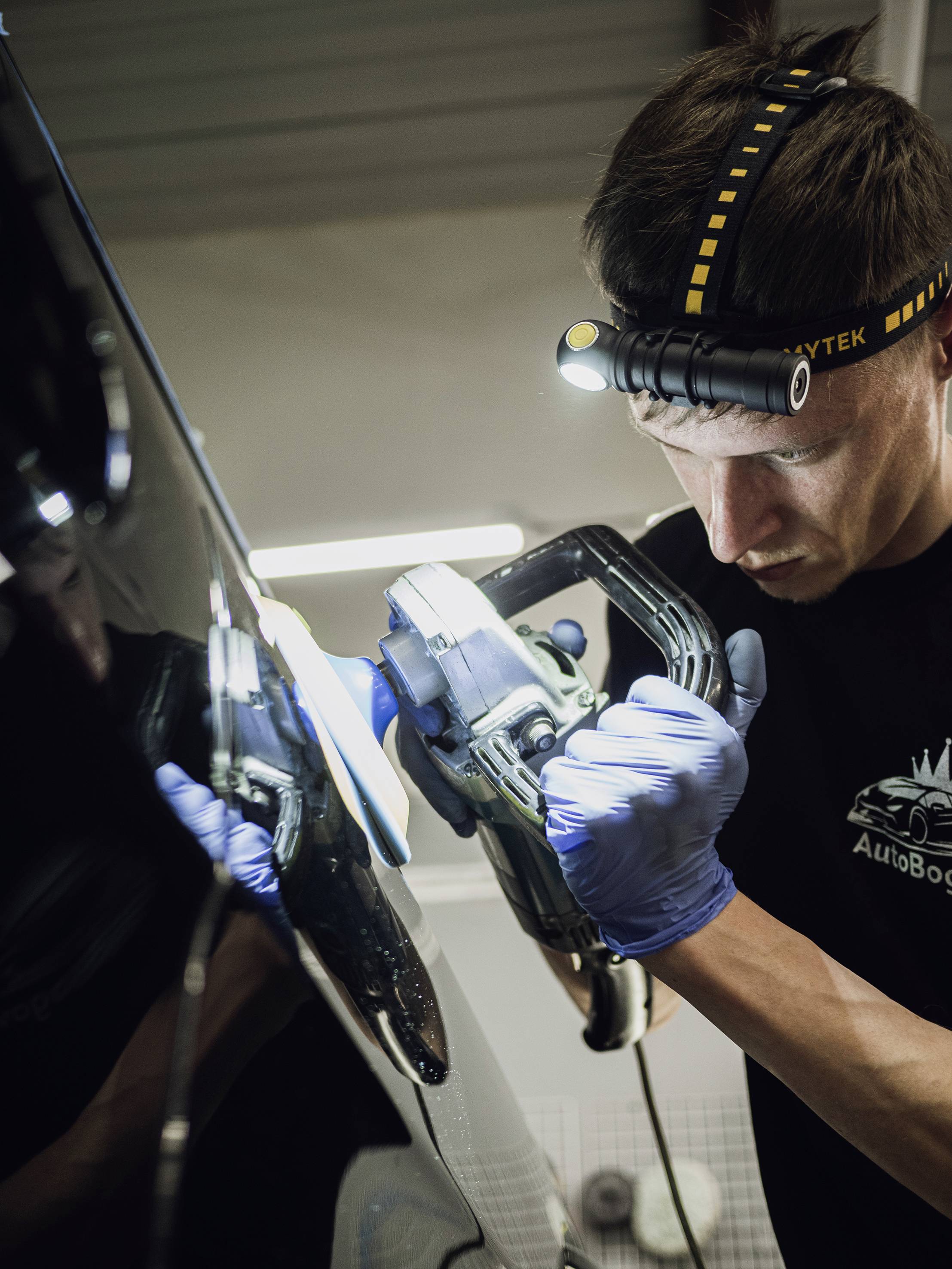 A mechanic is polishing a car with a polishing machine in a garage. He is wearing gloves and a head torch.