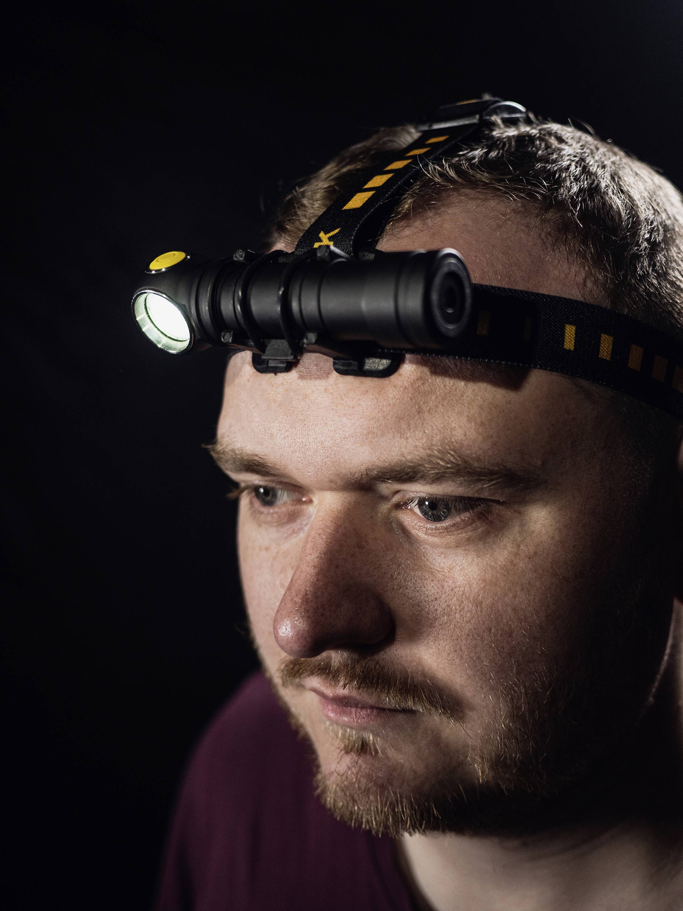 A man is wearing a head torch in a dark room. He is looking straight ahead, the head torch illuminating the space.