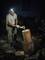 A person is splitting wood with an axe at night. They are wearing a head torch and camouflage trousers. Woodpiles can be seen in the background.