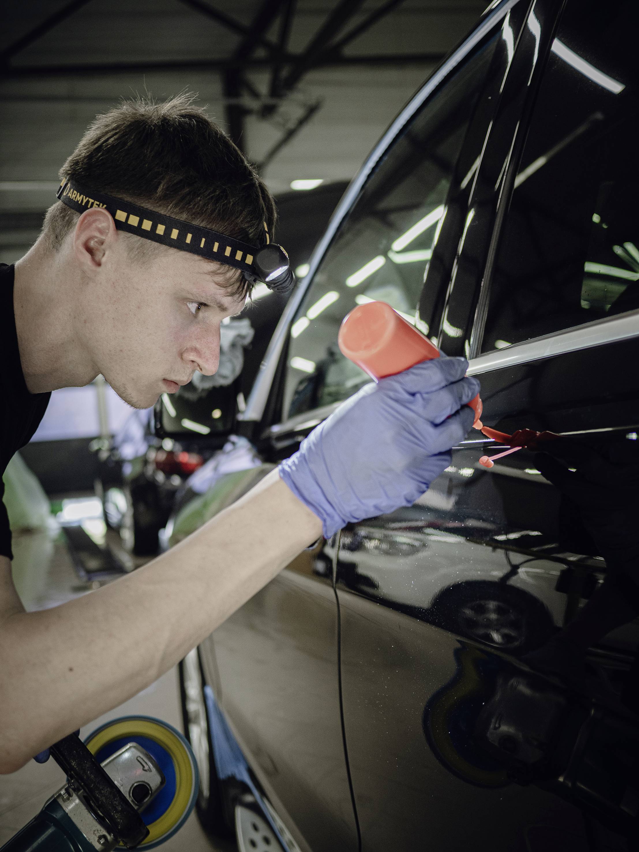 A man wearing a head torch and gloves carefully polishes the paintwork of a car with a polishing machine in a workshop.