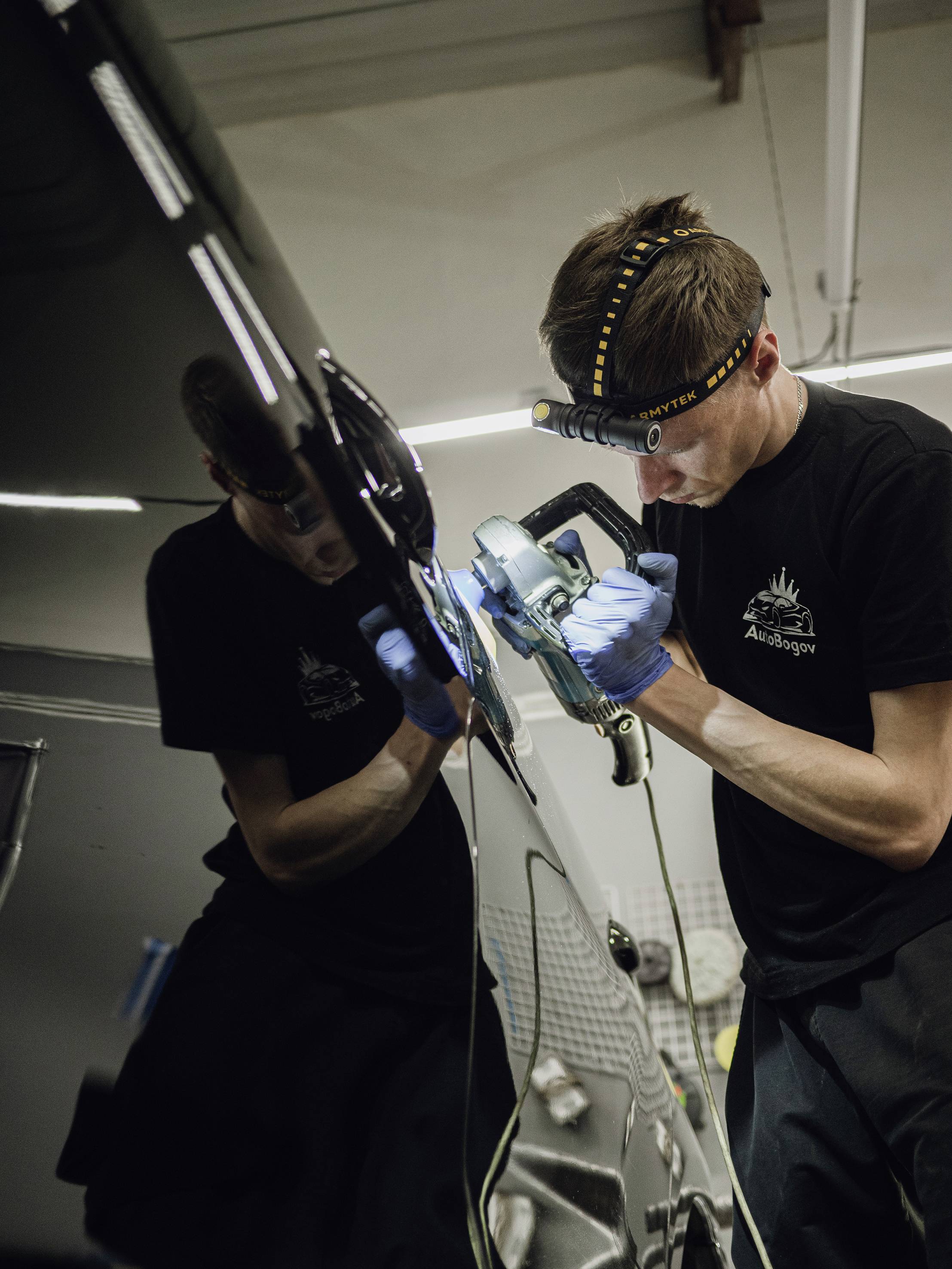 A person is polishing a car, wearing gloves and a head torch, while working with a machine, reflected on the car door.