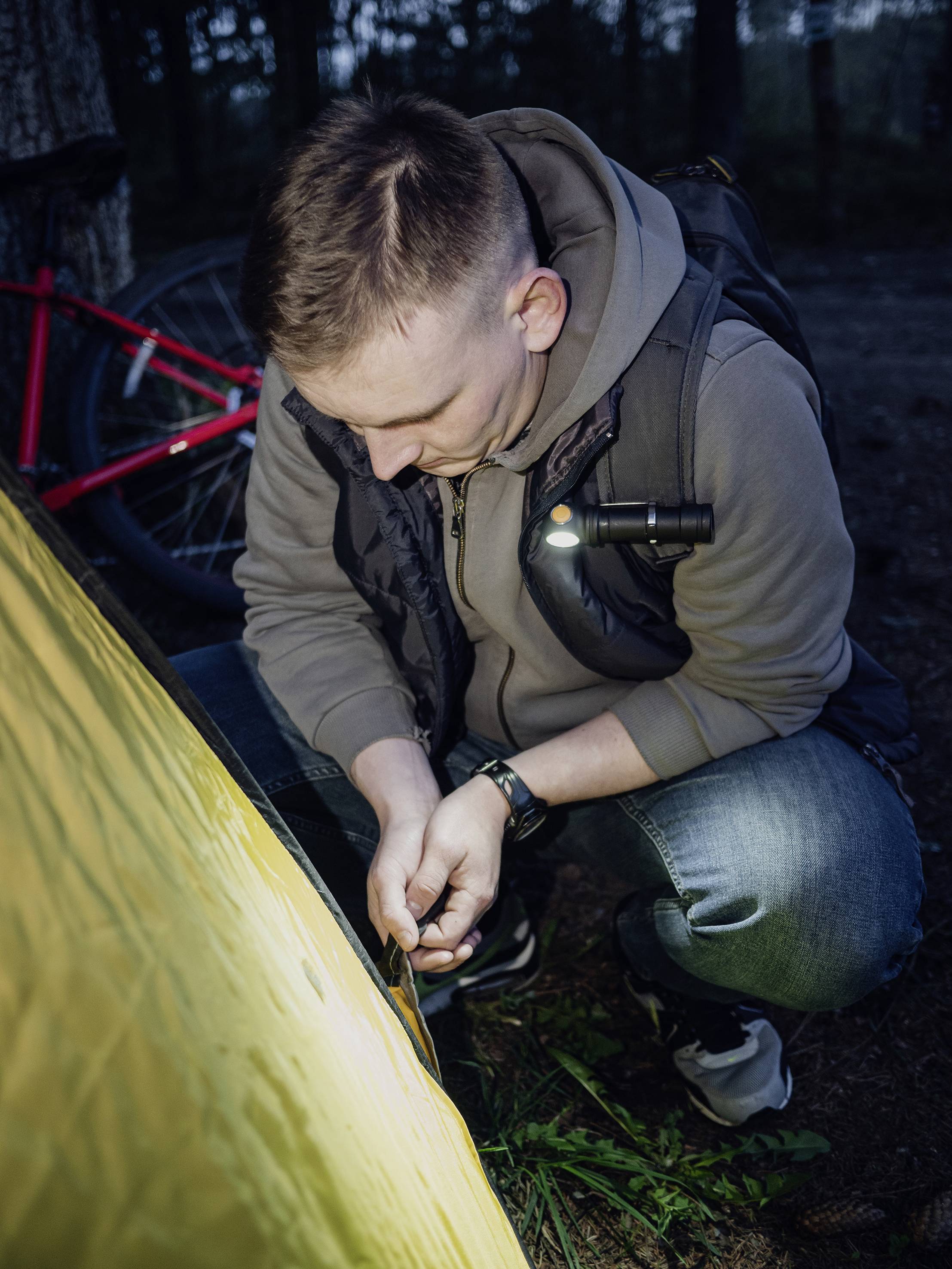 A man is kneeling on the ground at dusk, securing a tent. A red bicycle can be seen in the background.